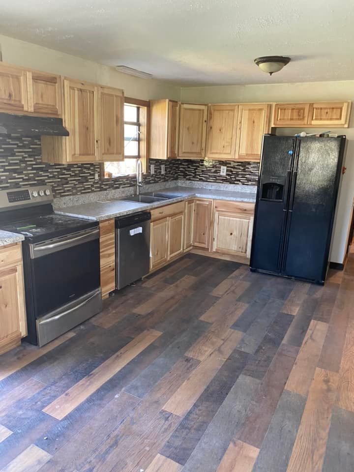 A kitchen with light wood cabinets, dark wood-look plank flooring, stainless steel stove, dishwasher, and black fridge.