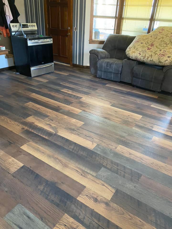 A living room featuring multi-toned wood plank flooring, a black oven, and a grey recliner couch by a window.