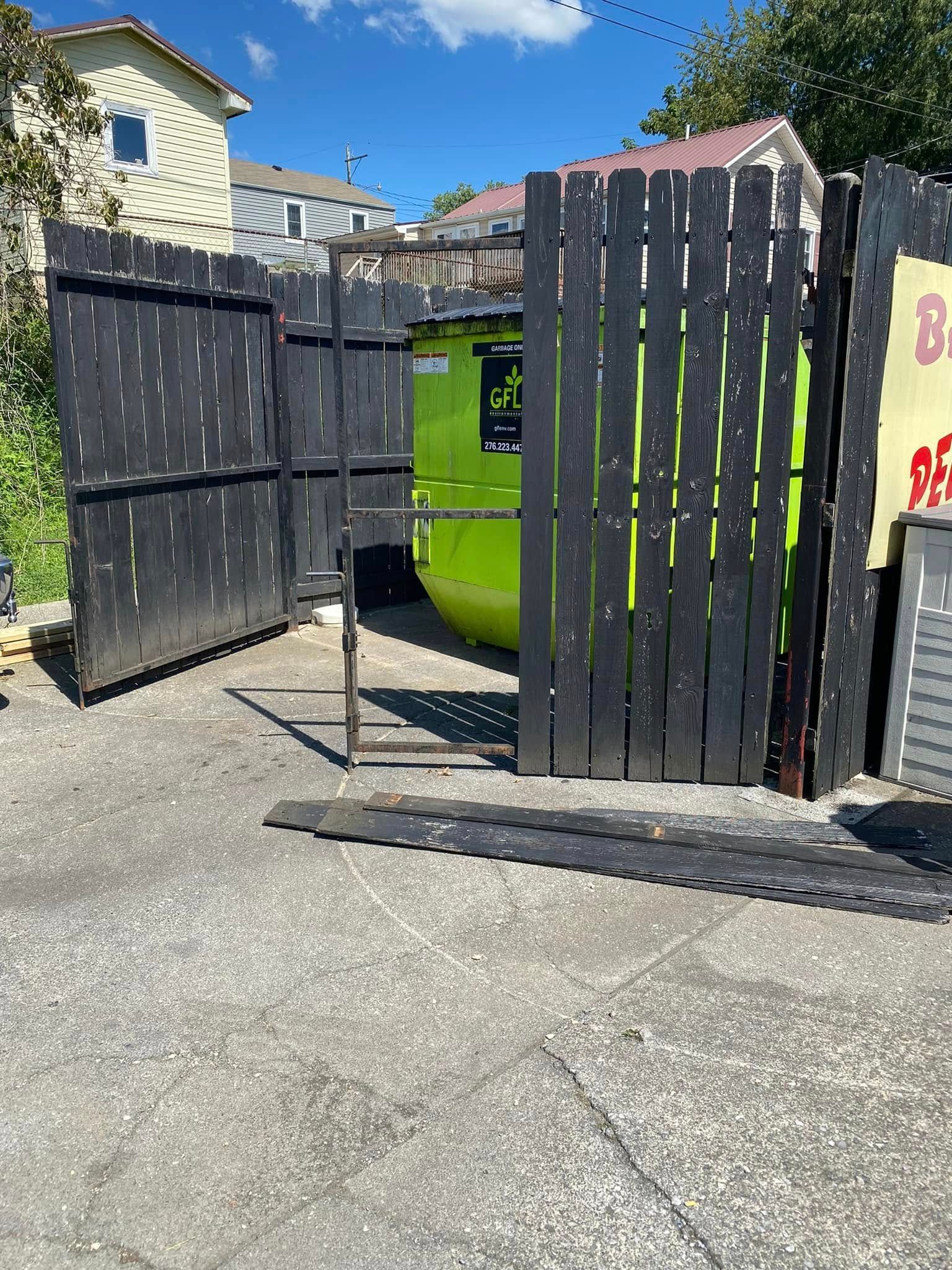 A bright green dumpster is partially hidden by a black wooden fence in a paved outdoor area.