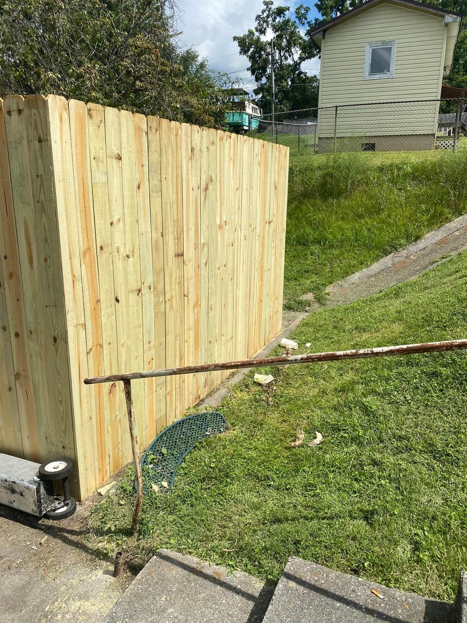 A newly constructed light-wood fence stands on a grassy slope next to a set of concrete steps and a residential building.