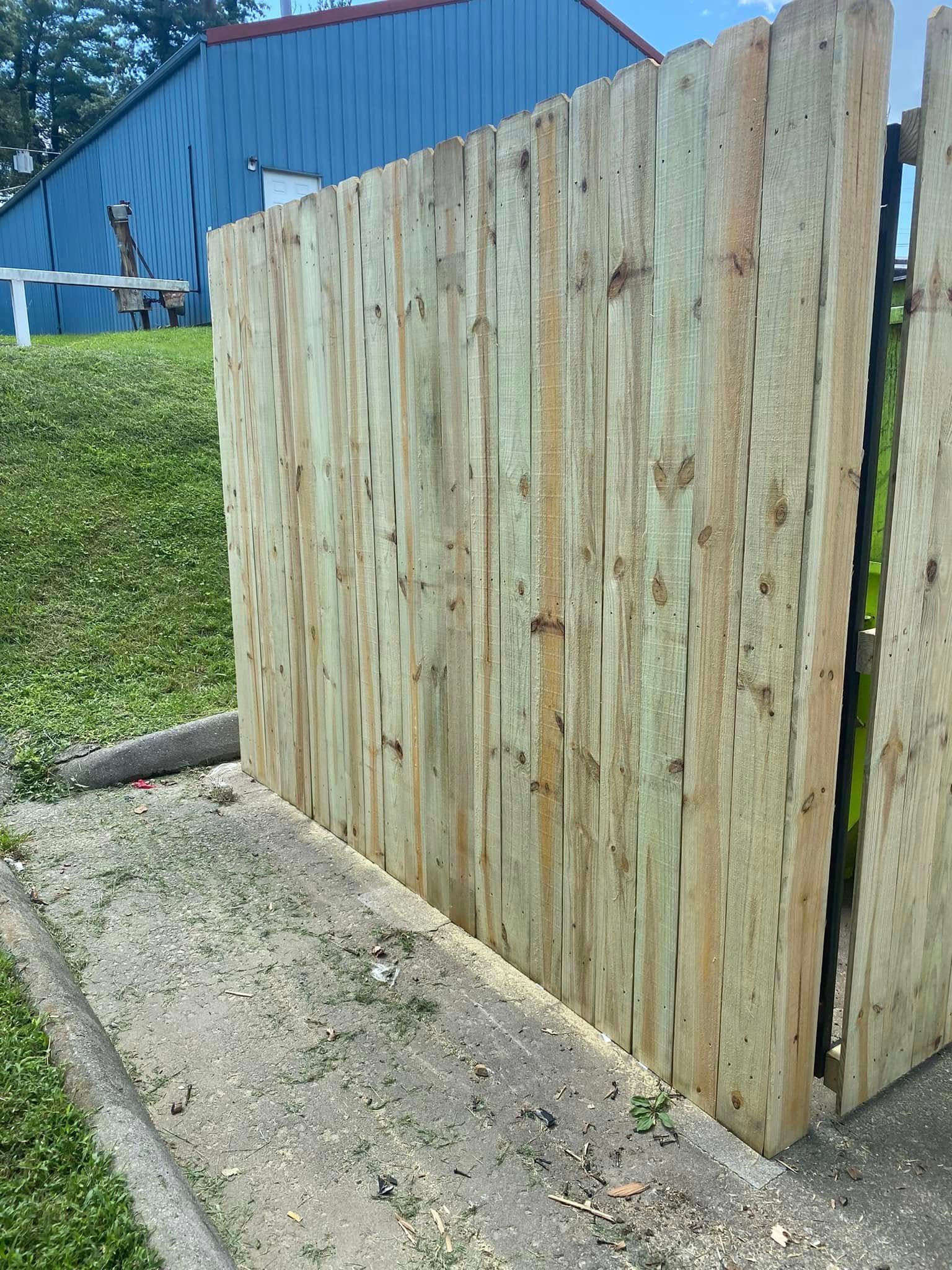 A side view of a newly installed light-colored wooden fence standing on a gravel patch near a blue building and grass.