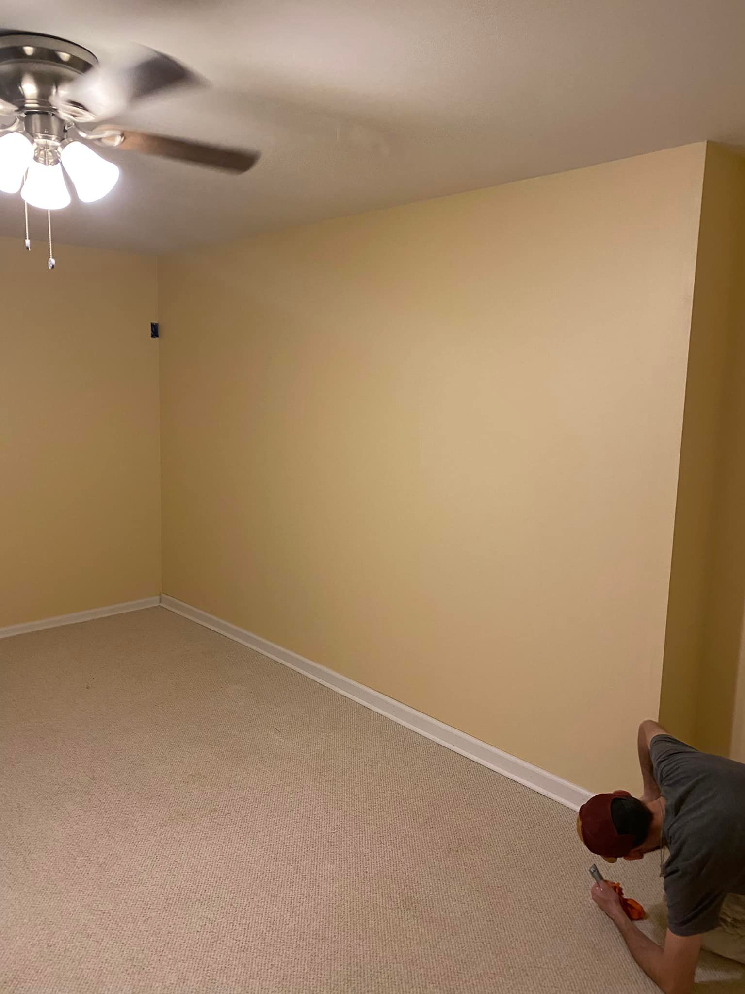 A person working on the carpeted floor in a room with light beige walls and a ceiling fan.