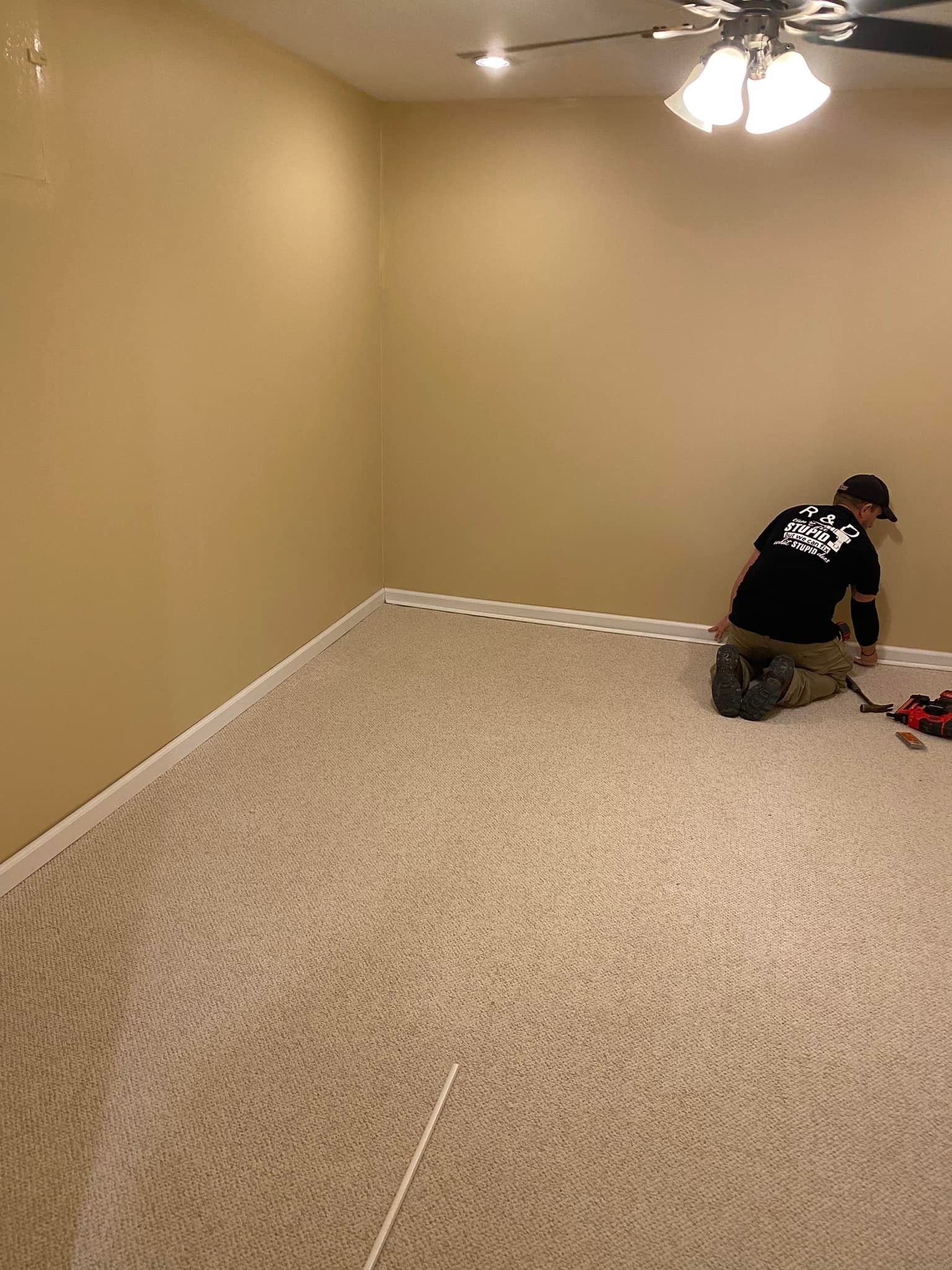 A person in a black shirt installs light-colored carpet in an empty, tan-walled room with a ceiling fan.