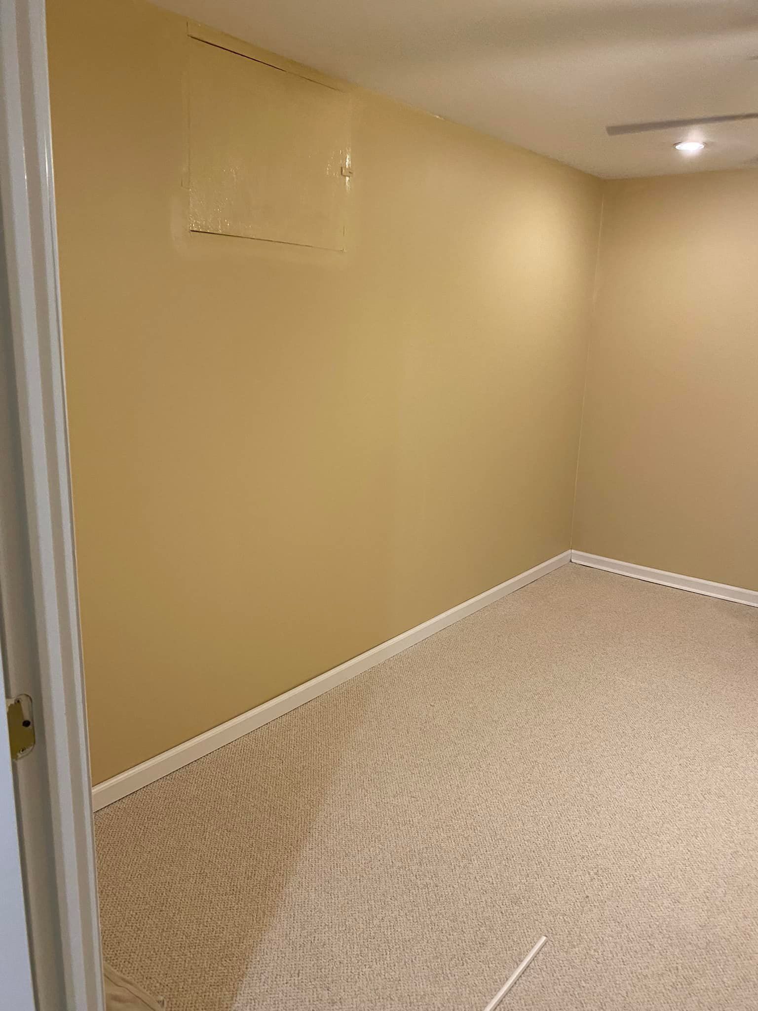 An empty room with tan walls, carpeted floors, and a small square access panel on the upper wall.