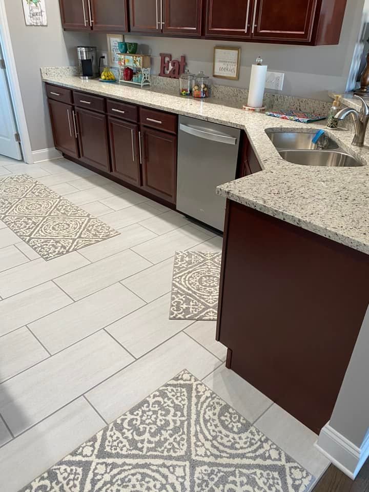 A kitchen with dark wood cabinets, light granite countertops, a stainless steel dishwasher, and patterned floor rugs.