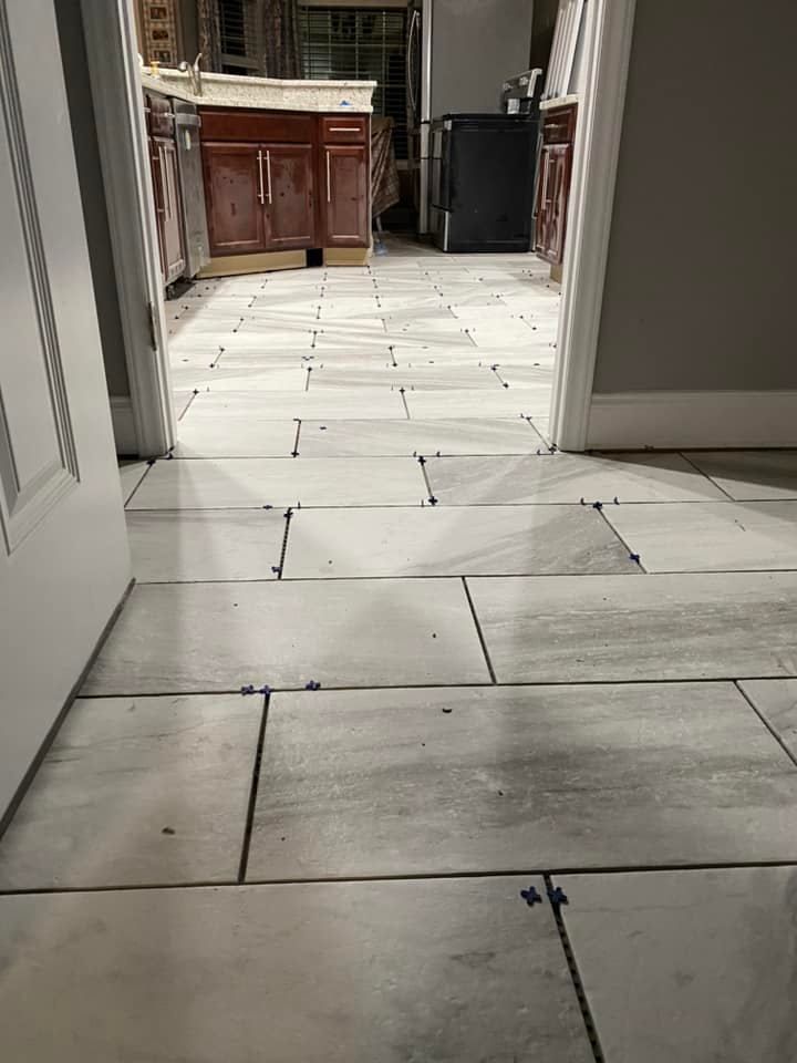 A view through a doorway into a kitchen featuring light grey, rectangular floor tiles currently being installed.