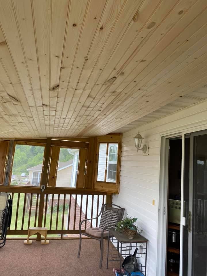 A wooden-paneled porch ceiling above a deck with a railing, a chair, a small table, and a glass sliding door.