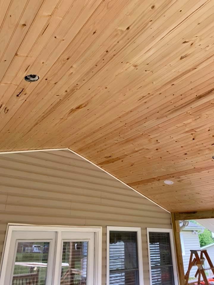 A low-angle view of a wooden beadboard ceiling on a covered porch, featuring recessed lighting and tan siding below.