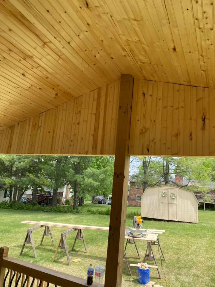 A wooden porch ceiling under construction, viewed from a deck overlooking a backyard with sawhorses and a shed.