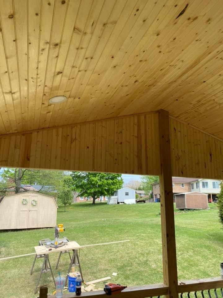 A covered wooden porch ceiling under construction overlooking a green backyard with a storage shed and a miter saw.