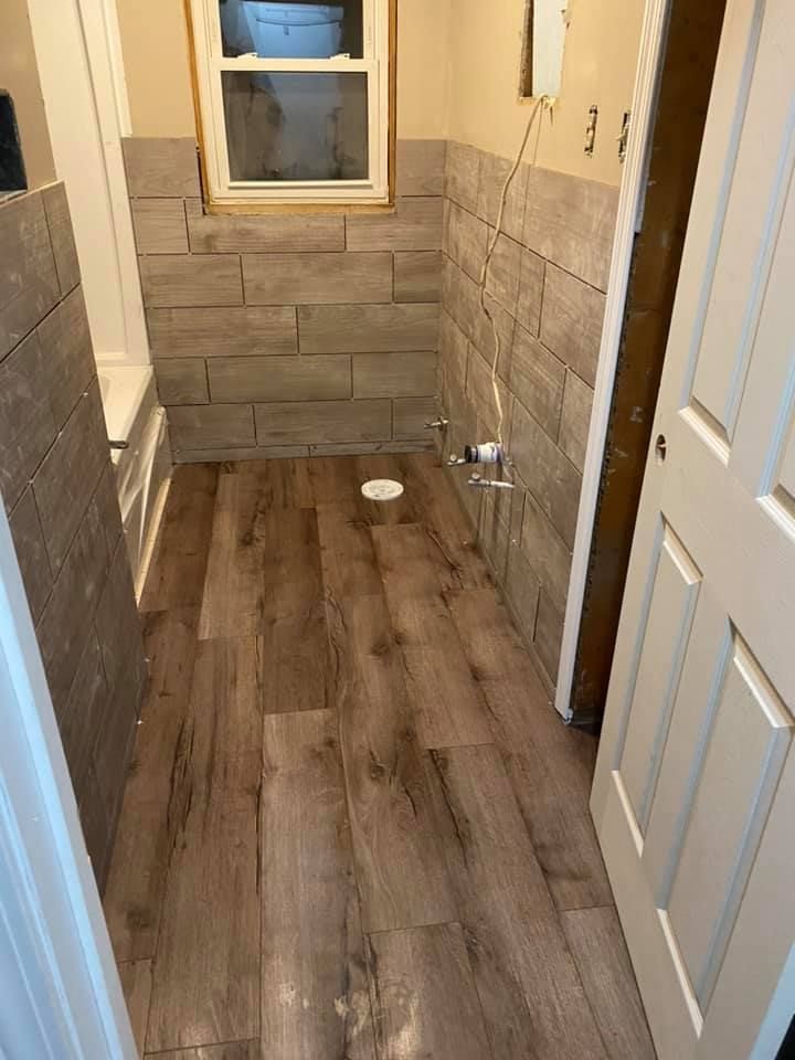 A bathroom under renovation with wood-look tile flooring and wainscoting, featuring a window and plumbing fixtures.