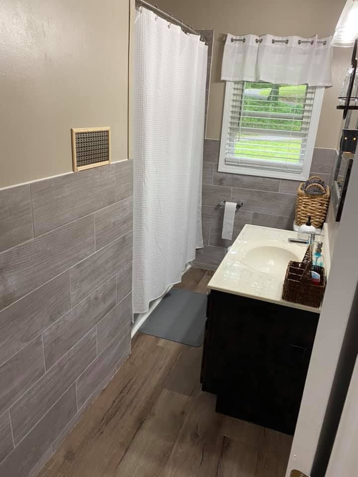 A bathroom interior with light wood-look floor tiles, grey wall tiles, a white shower curtain, and a dark wood vanity.