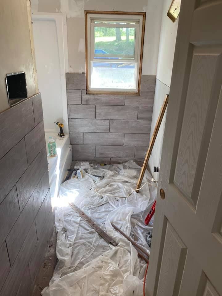 A bathroom under renovation with light gray wood-look wall tiles, a window, a bathtub, and drop cloths on the floor.