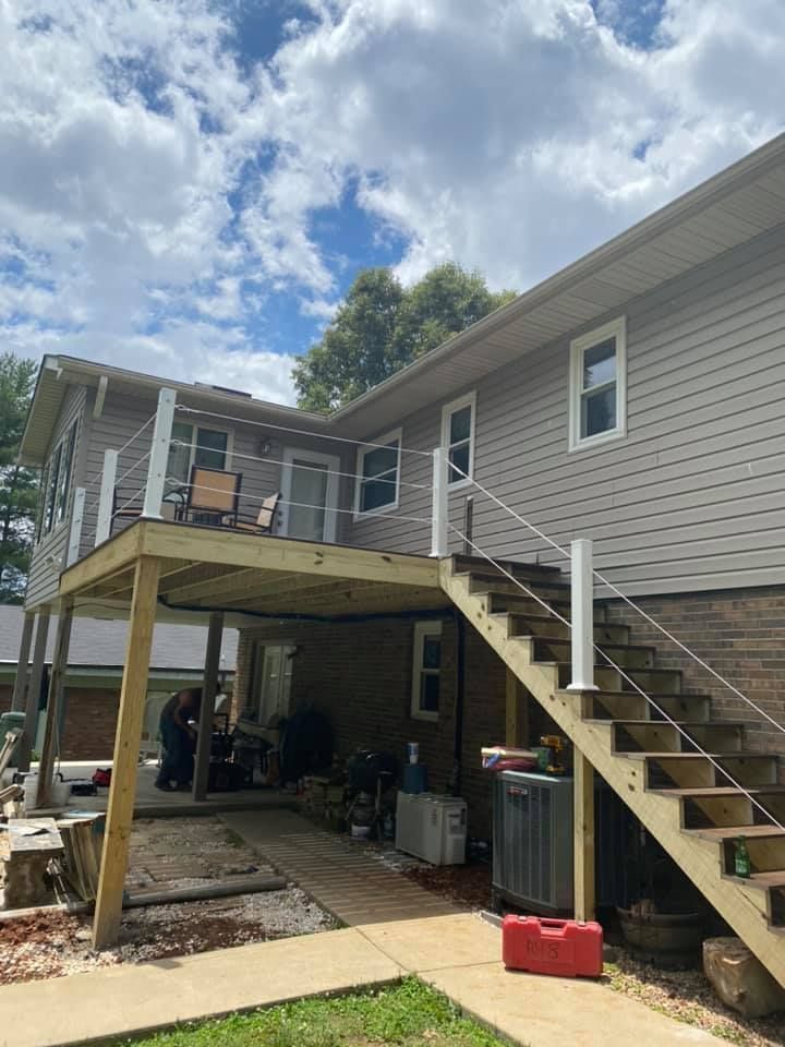 A wooden deck with cable railing and stairs leads up to a grey-sided house on a sunny day with a person working below.