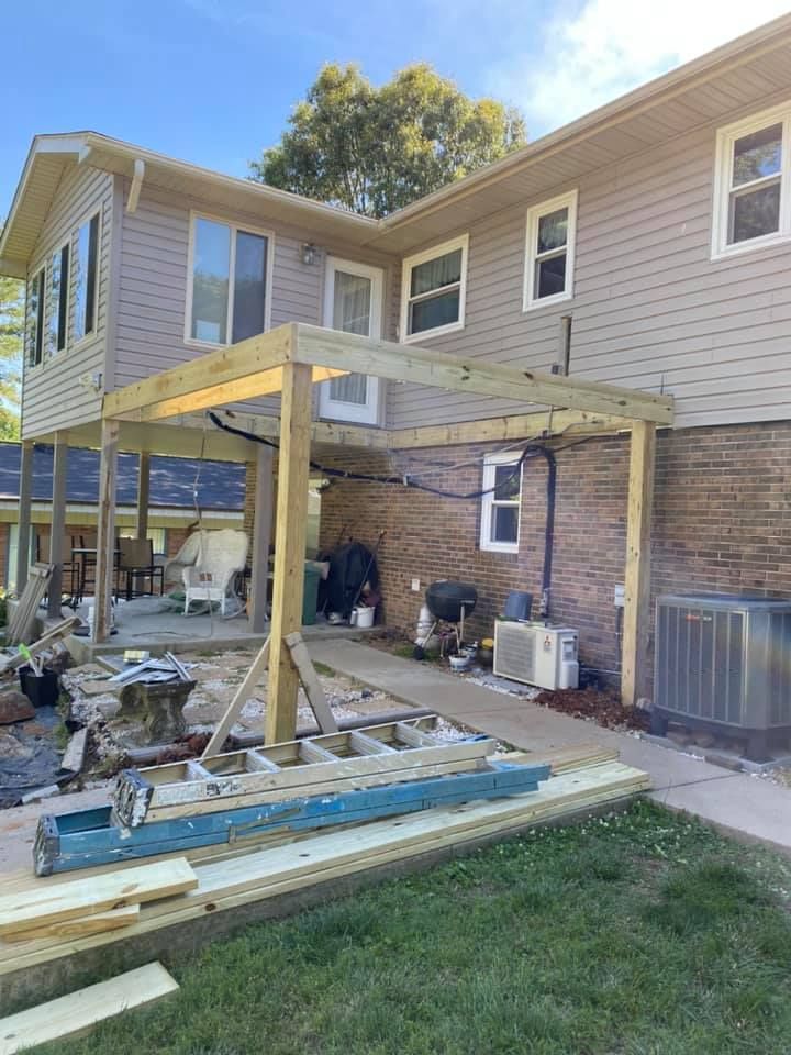 Construction of a wooden deck frame attached to a two-story house with tan siding and a brick lower level.