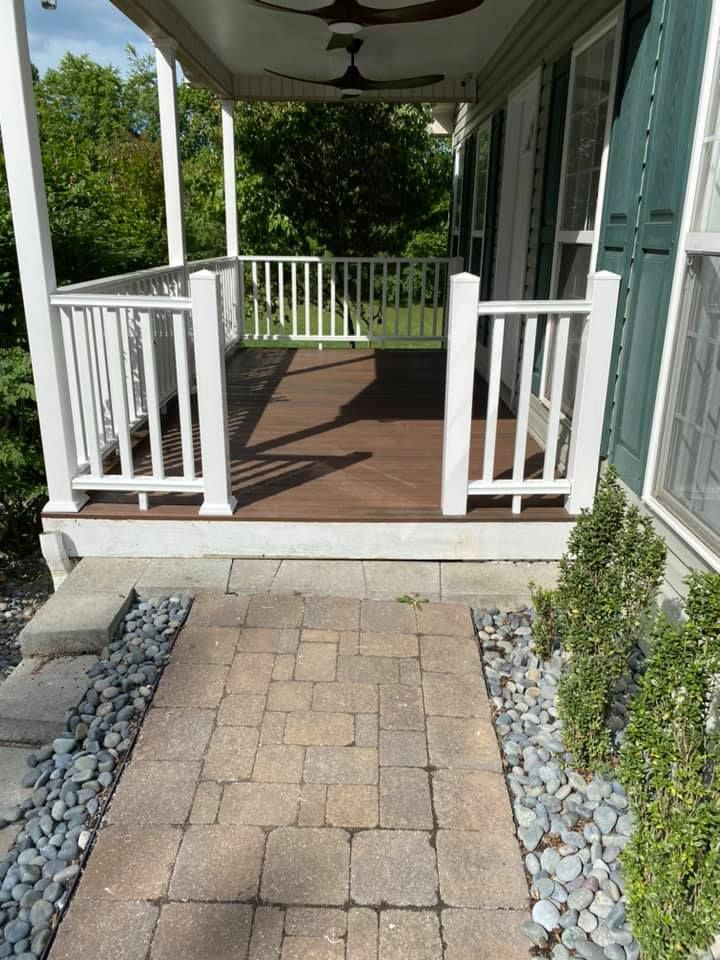 A covered wooden front porch with white railings, approached by a stone paver walkway bordered by grey stones and shrubs.