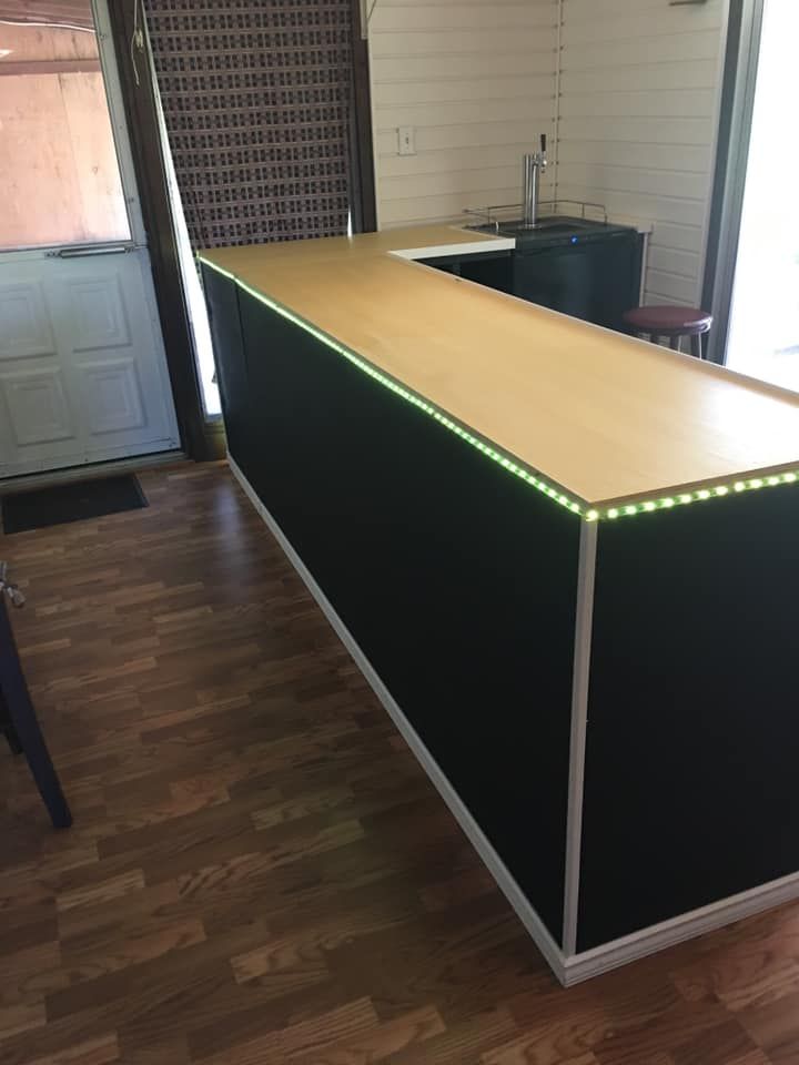 A black, L-shaped home bar with a light wood countertop and an integrated strip of glowing green LED lights.