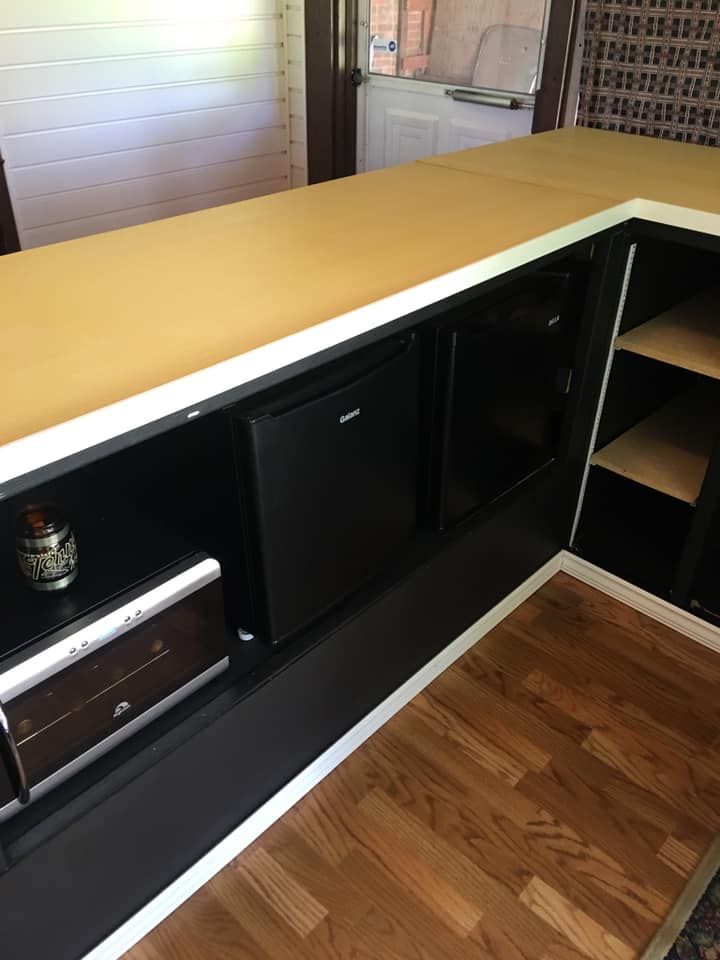 An L-shaped home bar counter with black cabinetry, a small refrigerator, and shelves, set on a wood-look floor.