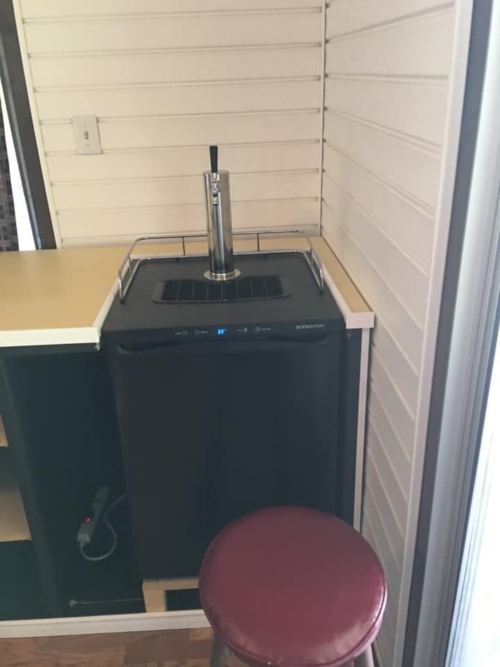 A black kegerator with a silver tap tower installed between white-paneled walls, with a red stool in the foreground.