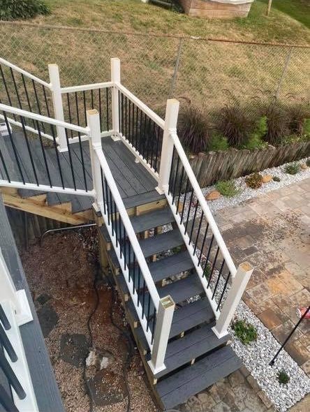 A high-angle view of outdoor gray composite stairs with white railings and black balusters, descending to a stone patio.