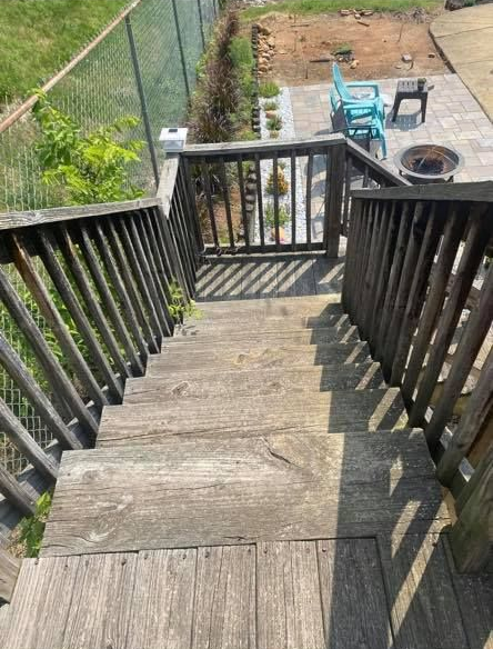 A high-angle view looking down wooden outdoor stairs toward a fenced yard with a small patio and seating area.