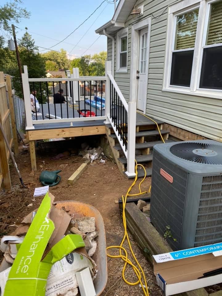 A wooden deck under construction with white railings, stairs, and construction debris in a backyard next to an AC unit.