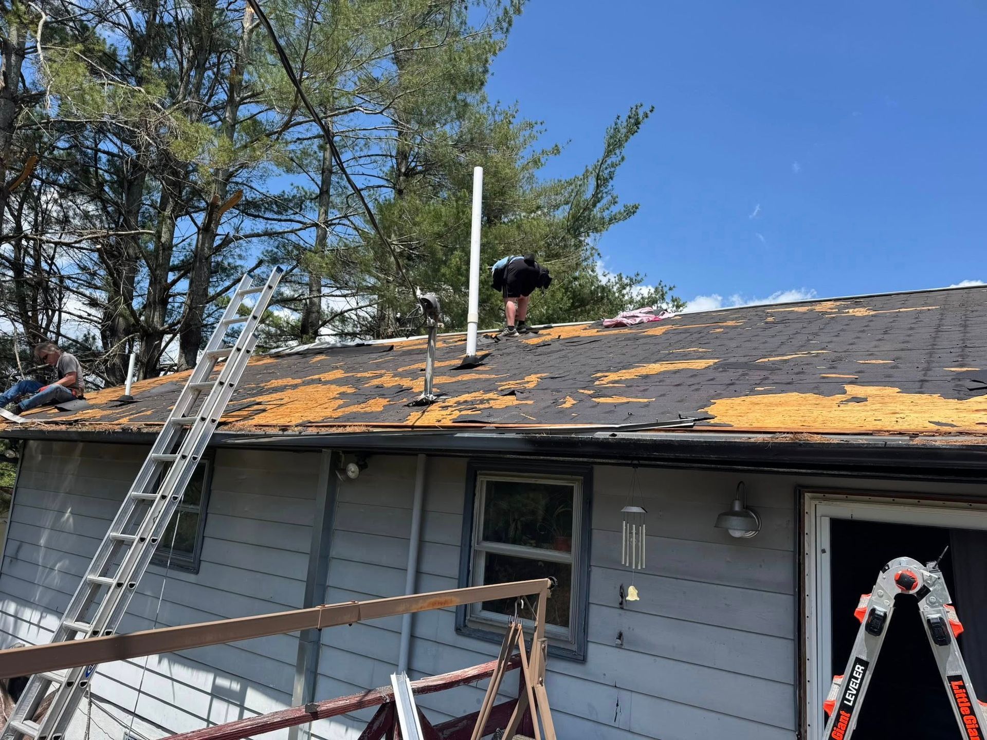 A before image of a roof with two individuals on it, working on a roofing project, with ladders and trees in the background.