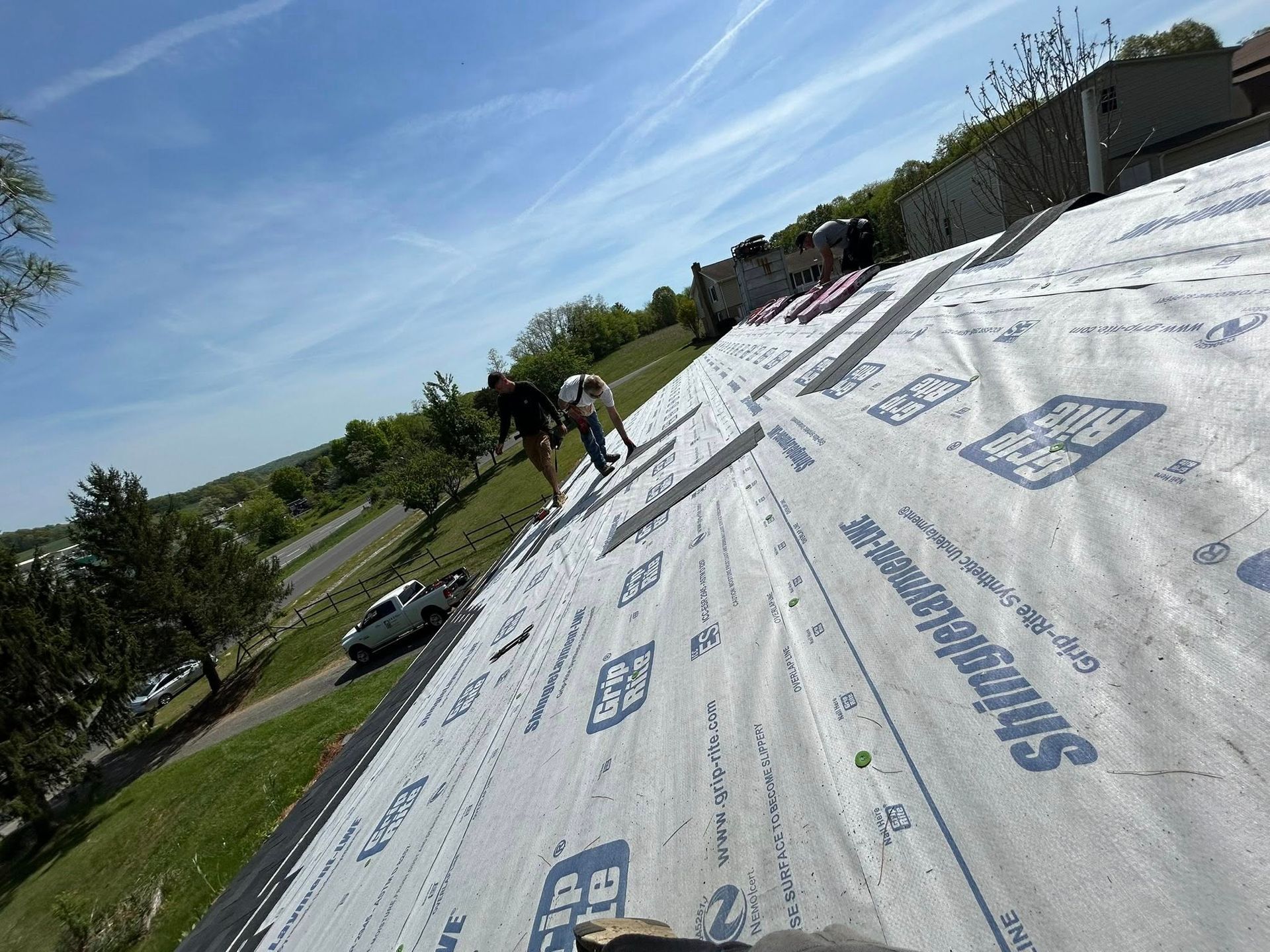 Workers install white protective underlayment on a sloping roof under a bright, sunny sky.