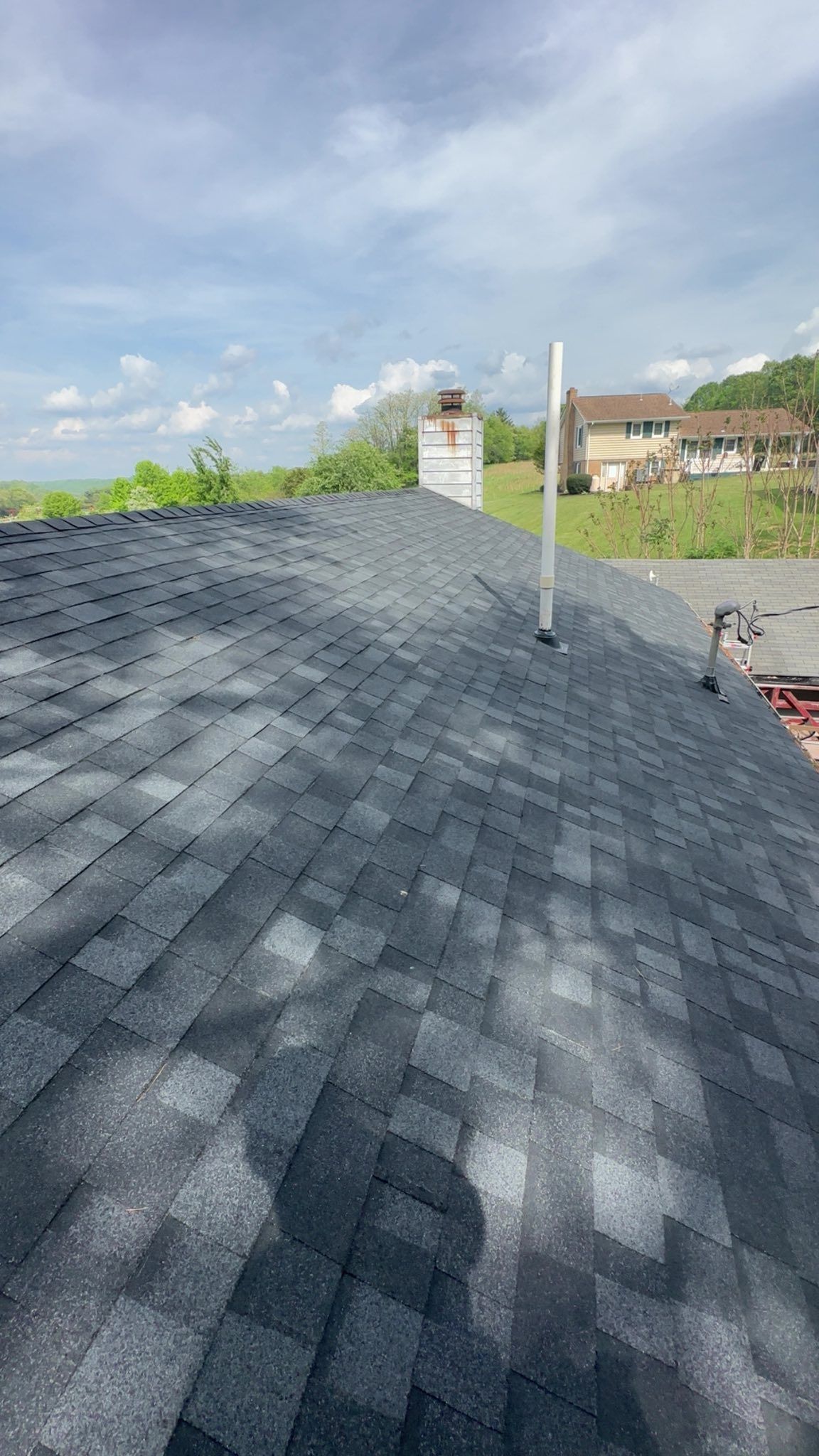 Gray asphalt shingles cover a sloped roof with a chimney, a ventilation pipe, and a partially visible house in the distance.