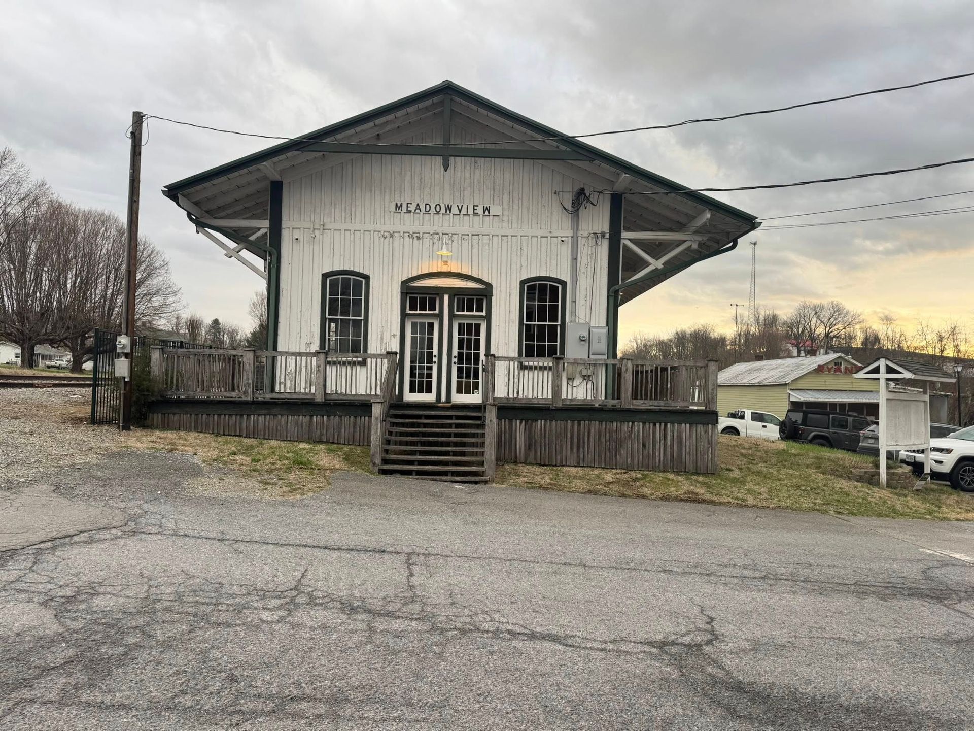 A historic white wooden train depot with a dark roof and stairs, set against a cloudy sky with nearby parked cars.