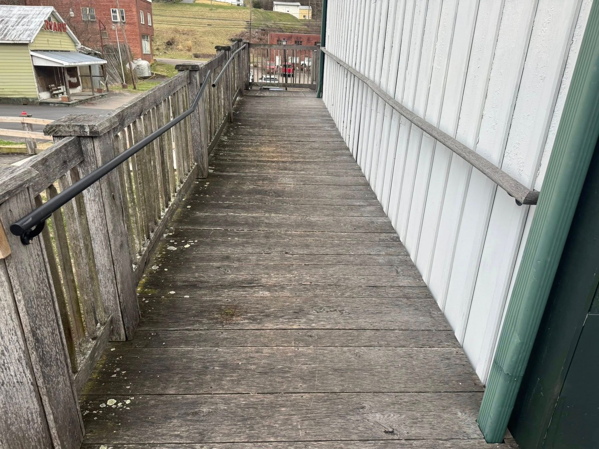 A before image of a weathered wooden walkway with handrails on both sides, leading toward a distant metal gate and buildings.