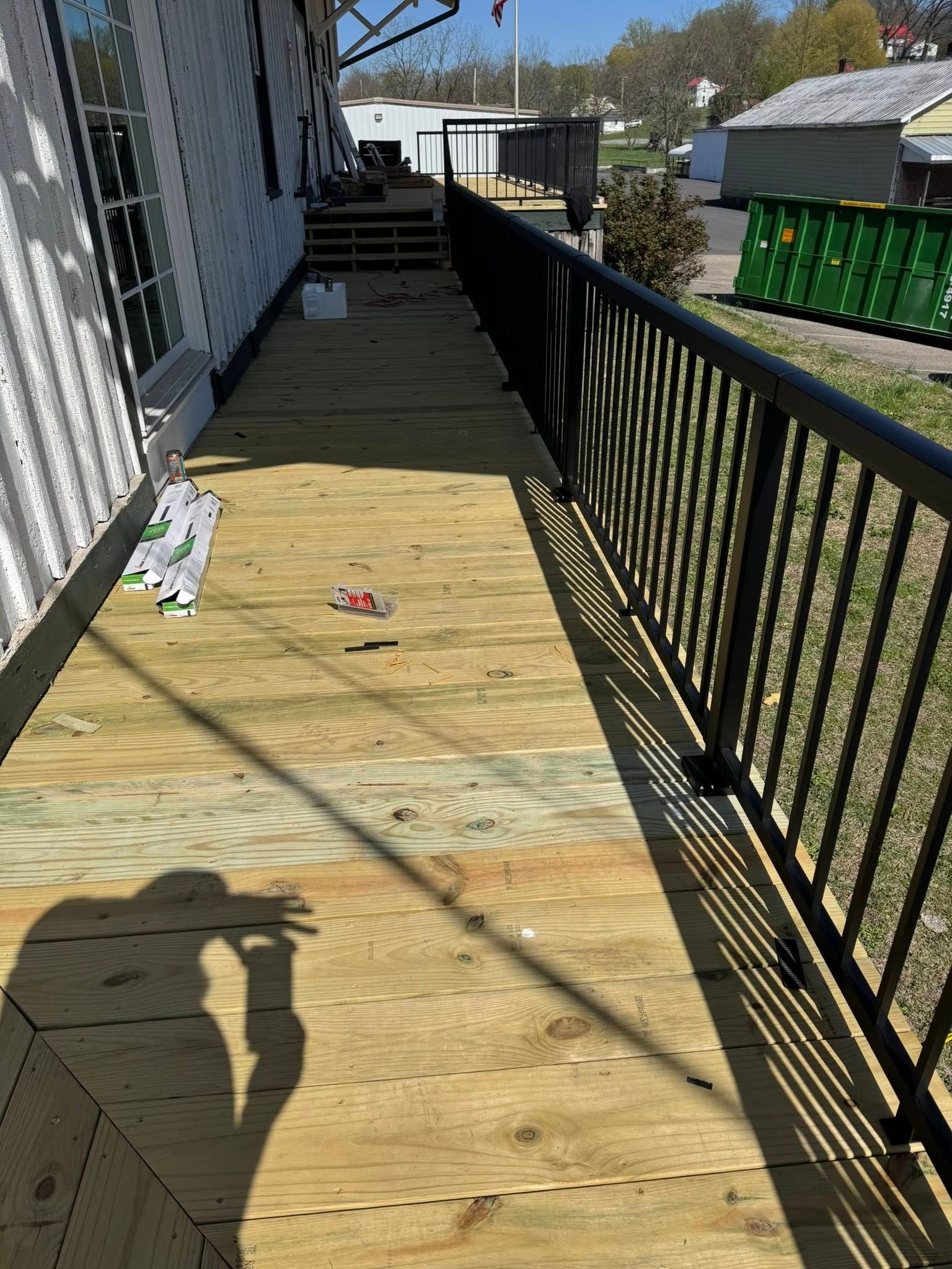 A newly installed wooden deck with a black metal railing runs along the side of a white building on a sunny day.