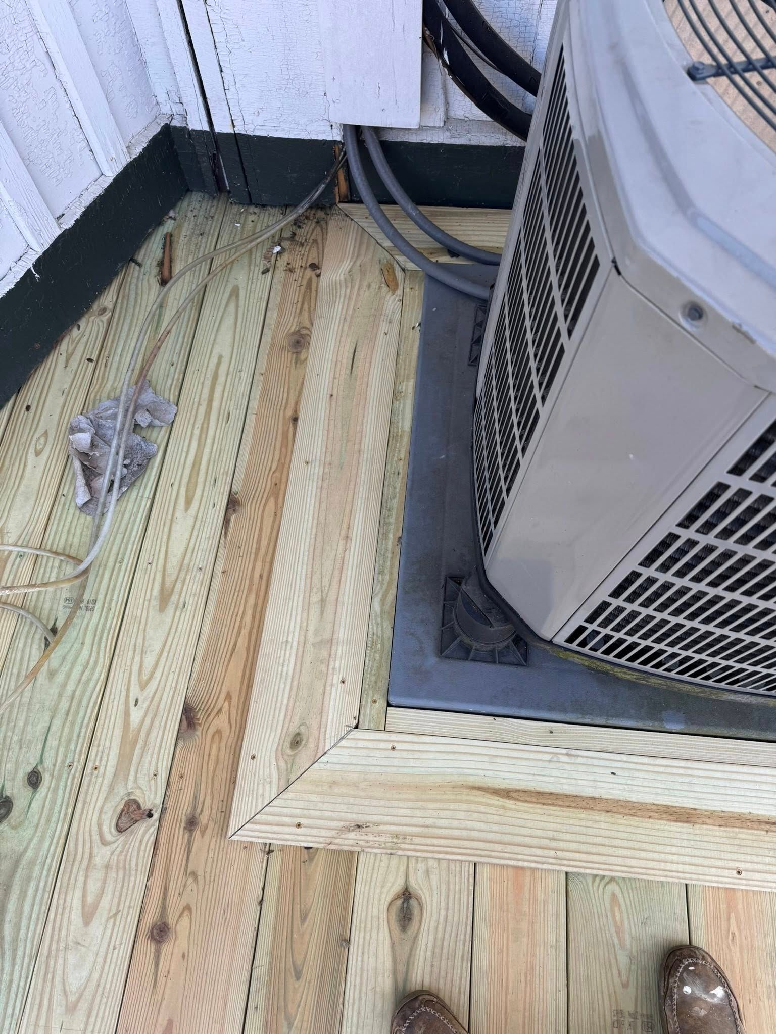 An outdoor air conditioning unit sits on a metal base surrounded by a newly built wooden deck frame.