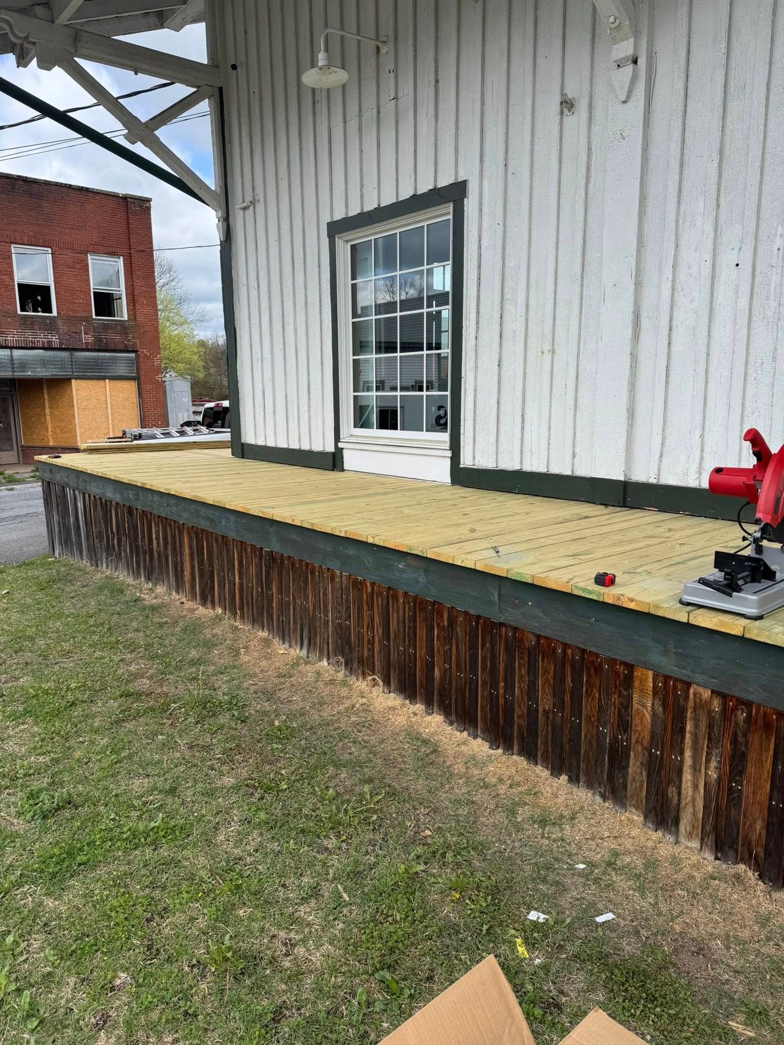 A red miter saw sits on a newly installed wooden deck platform beside a white building with vertical siding.
