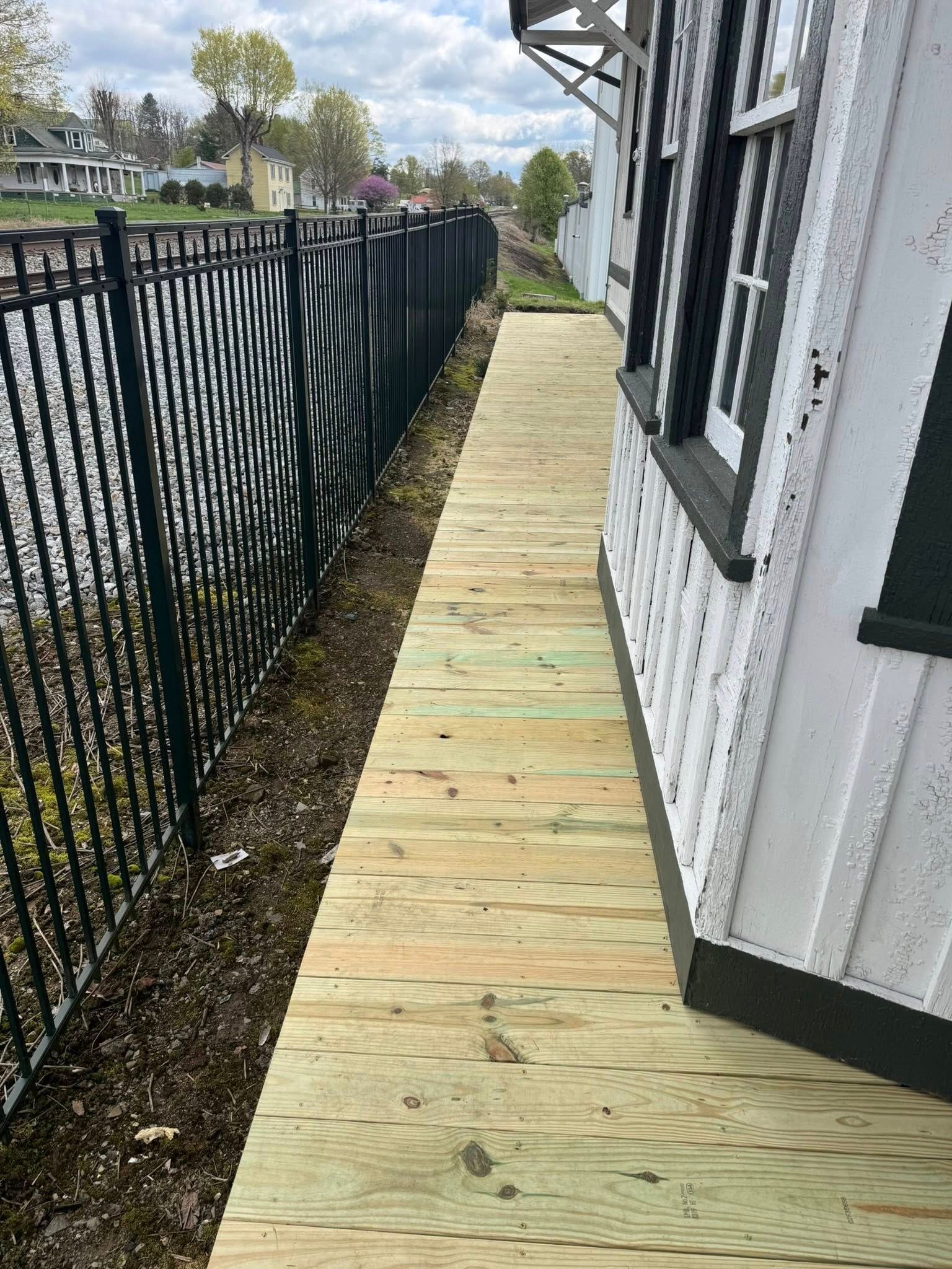 A narrow, newly installed wooden boardwalk runs along the side of a white building, adjacent to a black metal fence.