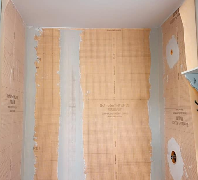 Shower wall installation showing beige Schluter-KERDI waterproof membrane over grey drywall with plumbing fixtures visible.