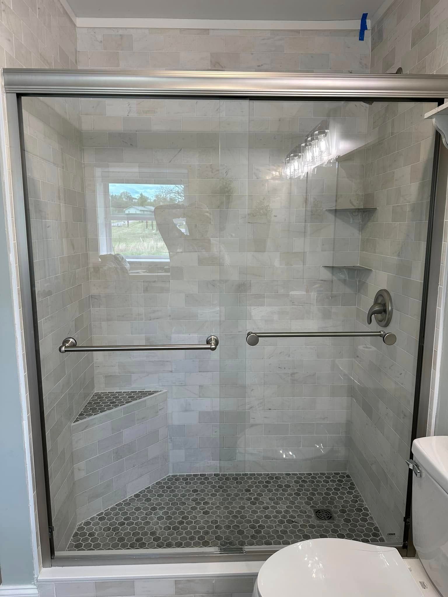 A walk-in shower with light grey tiled walls, a built-in corner bench, pebble floor, and glass sliding doors.