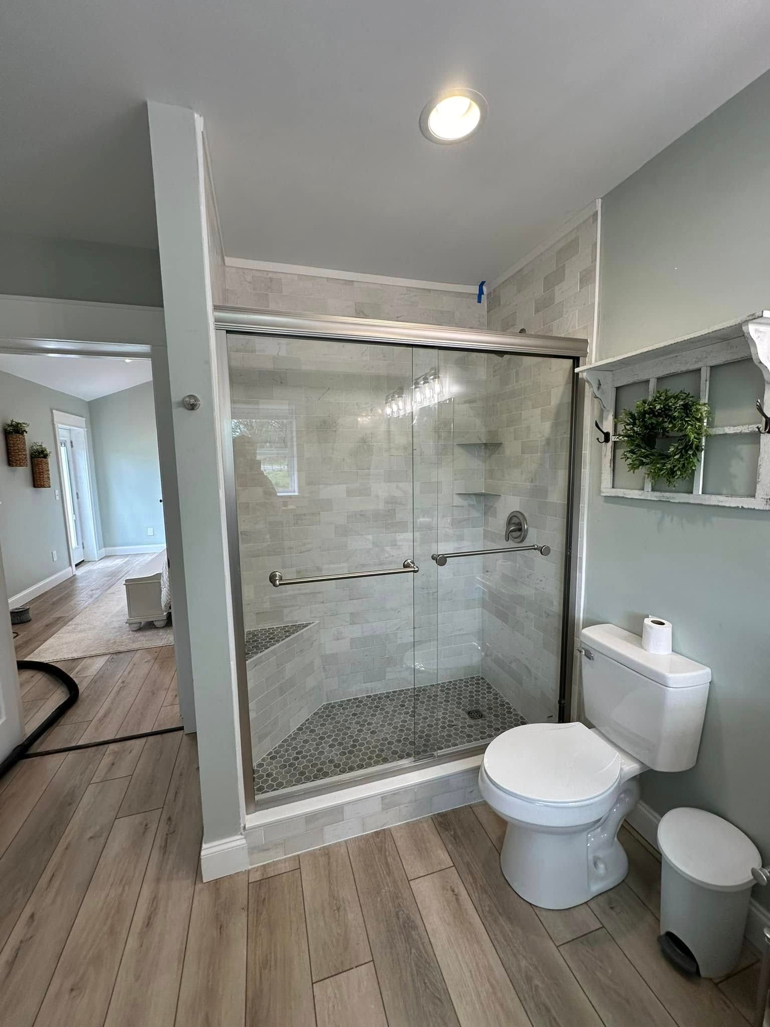A bathroom with light gray walls and wood-look flooring features a glass-enclosed shower with tiled walls and a toilet.