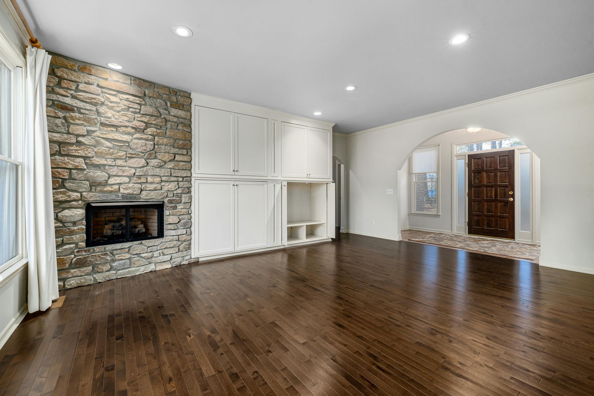 A living room with dark hardwood floors, a stone fireplace, white built-in cabinets, and an arched doorway.