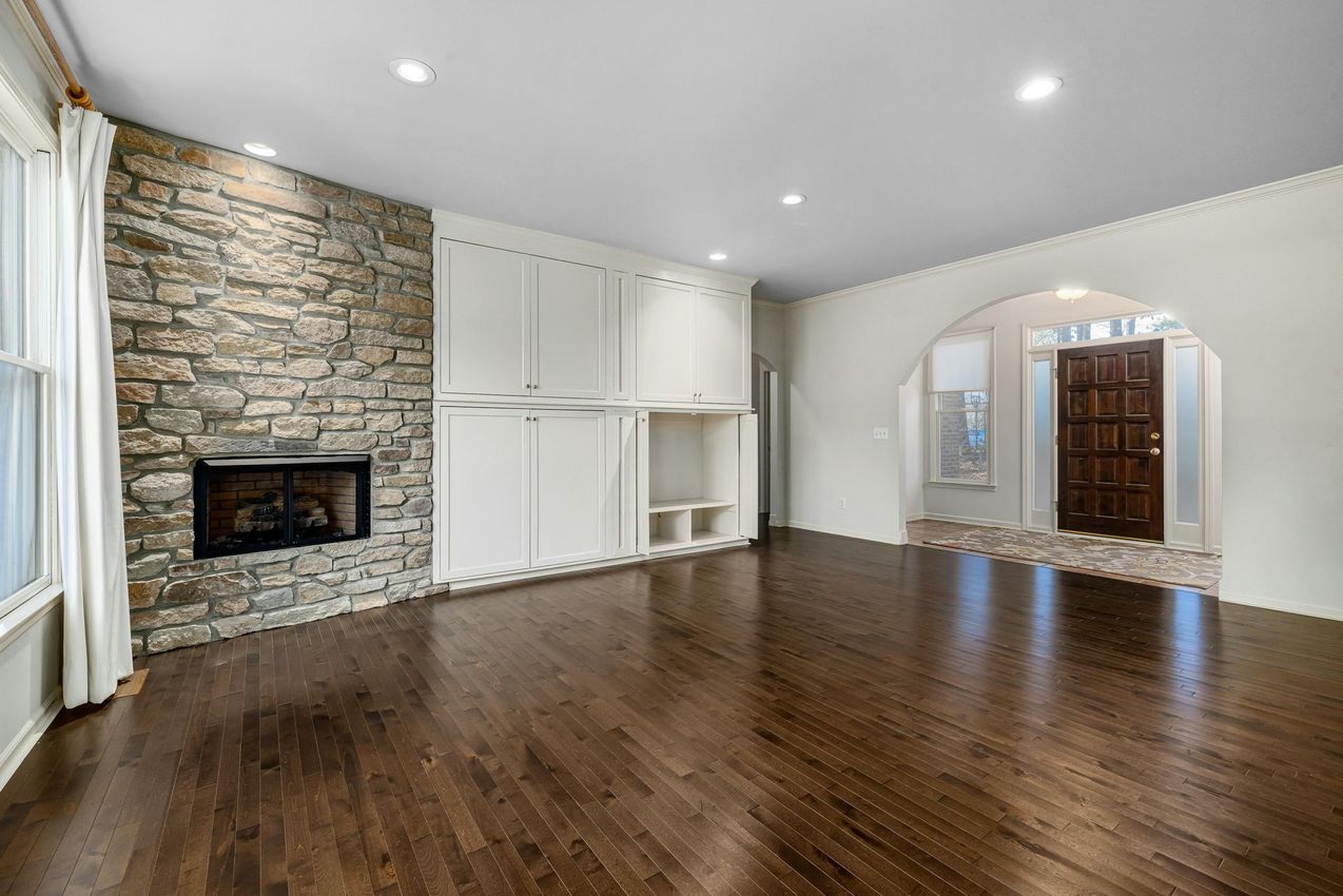 Contact Us A living room with dark hardwood floors, a stone fireplace, white built-in cabinets, and an arched entry to the foyer.
