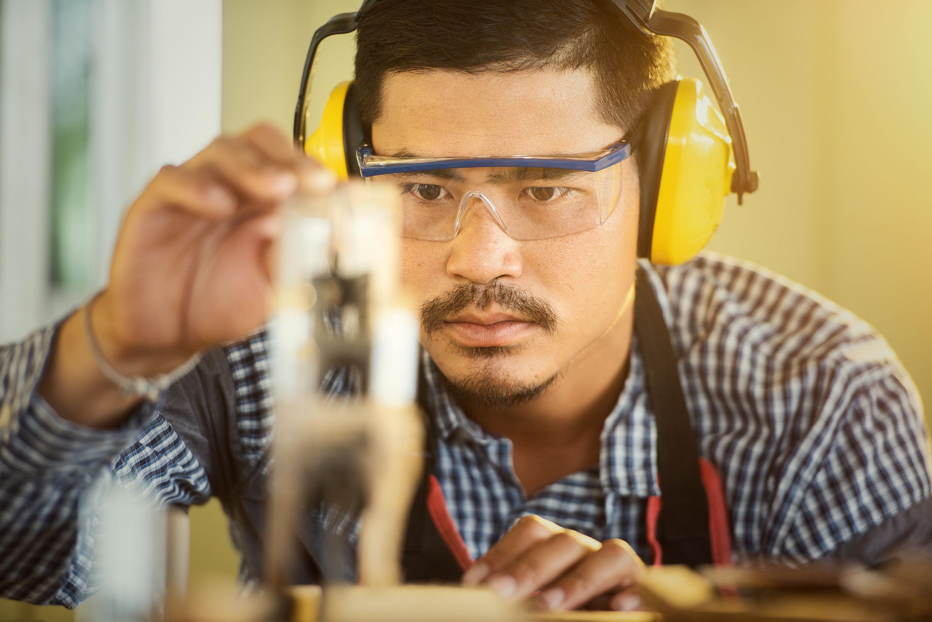 A craftsperson wearing safety glasses and ear protection intently inspects a small wooden project in a workshop.