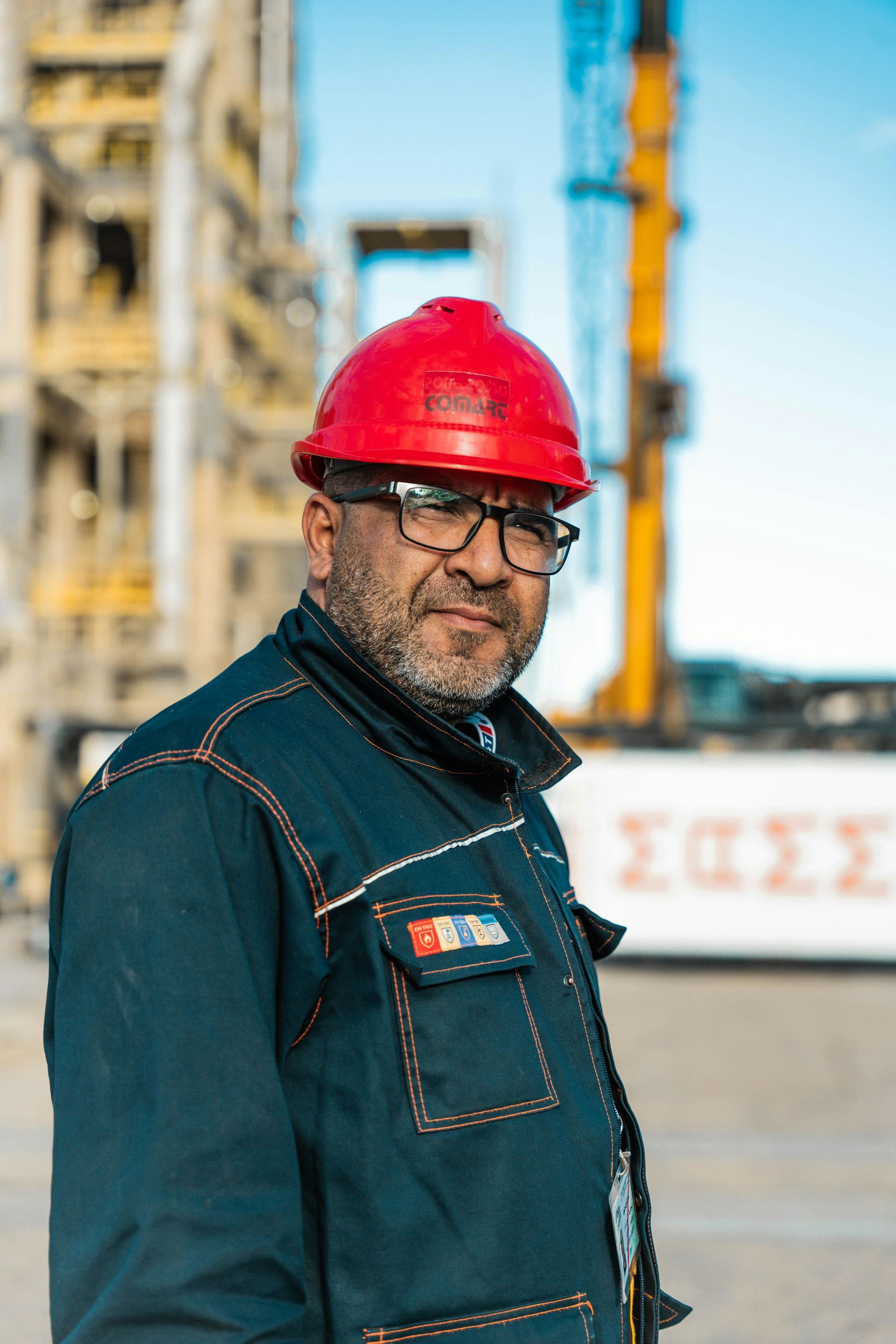 A person wearing a red hard hat and a dark blue work jacket stands in front of a construction site with a yellow crane.