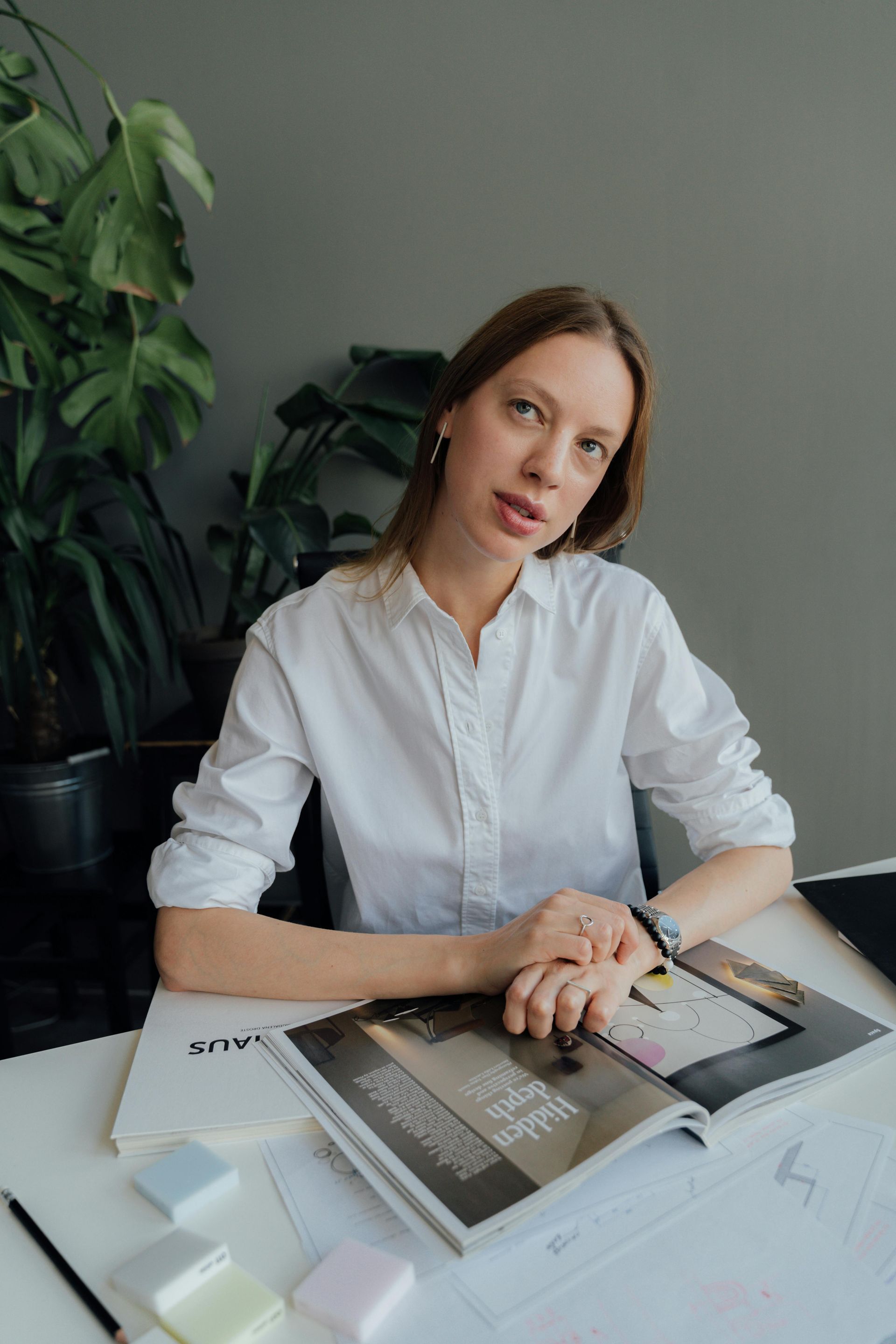 A person in a white button-down shirt sits at a desk with an open magazine, papers, and small decor items in a room.