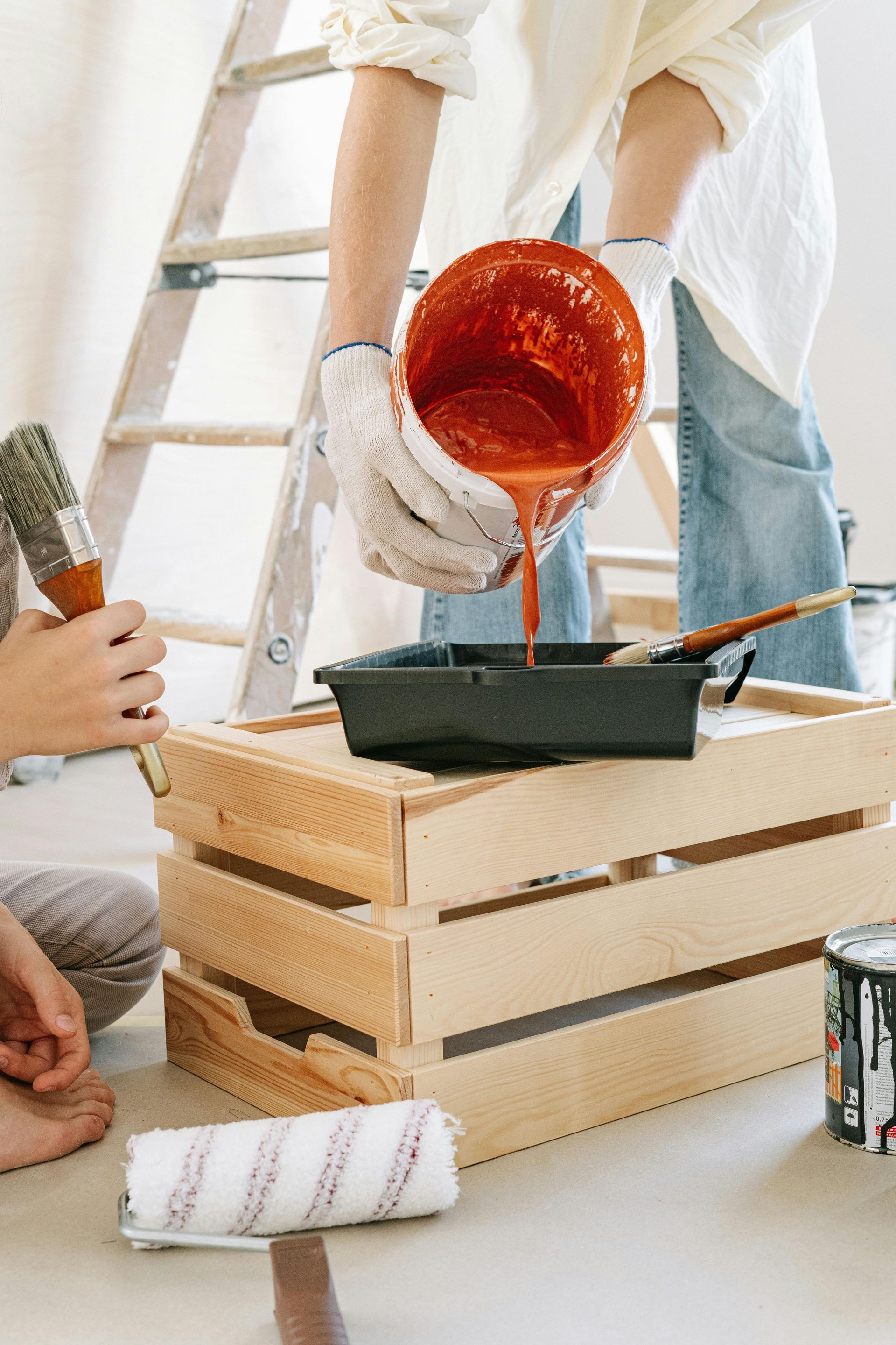 A person pouring terracotta-colored paint from a bucket into a tray resting on a wooden crate.