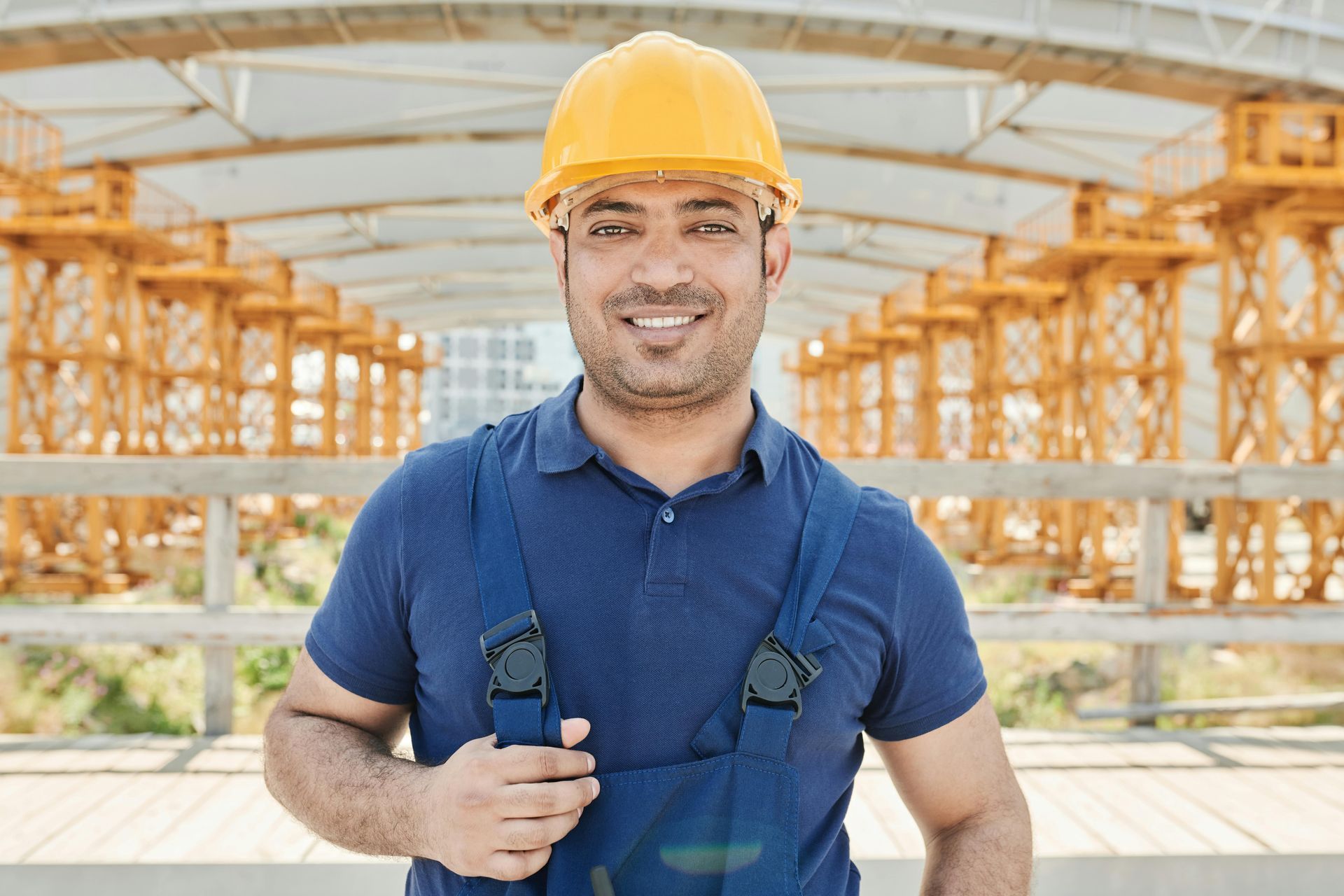 A smiling worker in a yellow hard hat and blue overalls standing in front of an outdoor construction site.