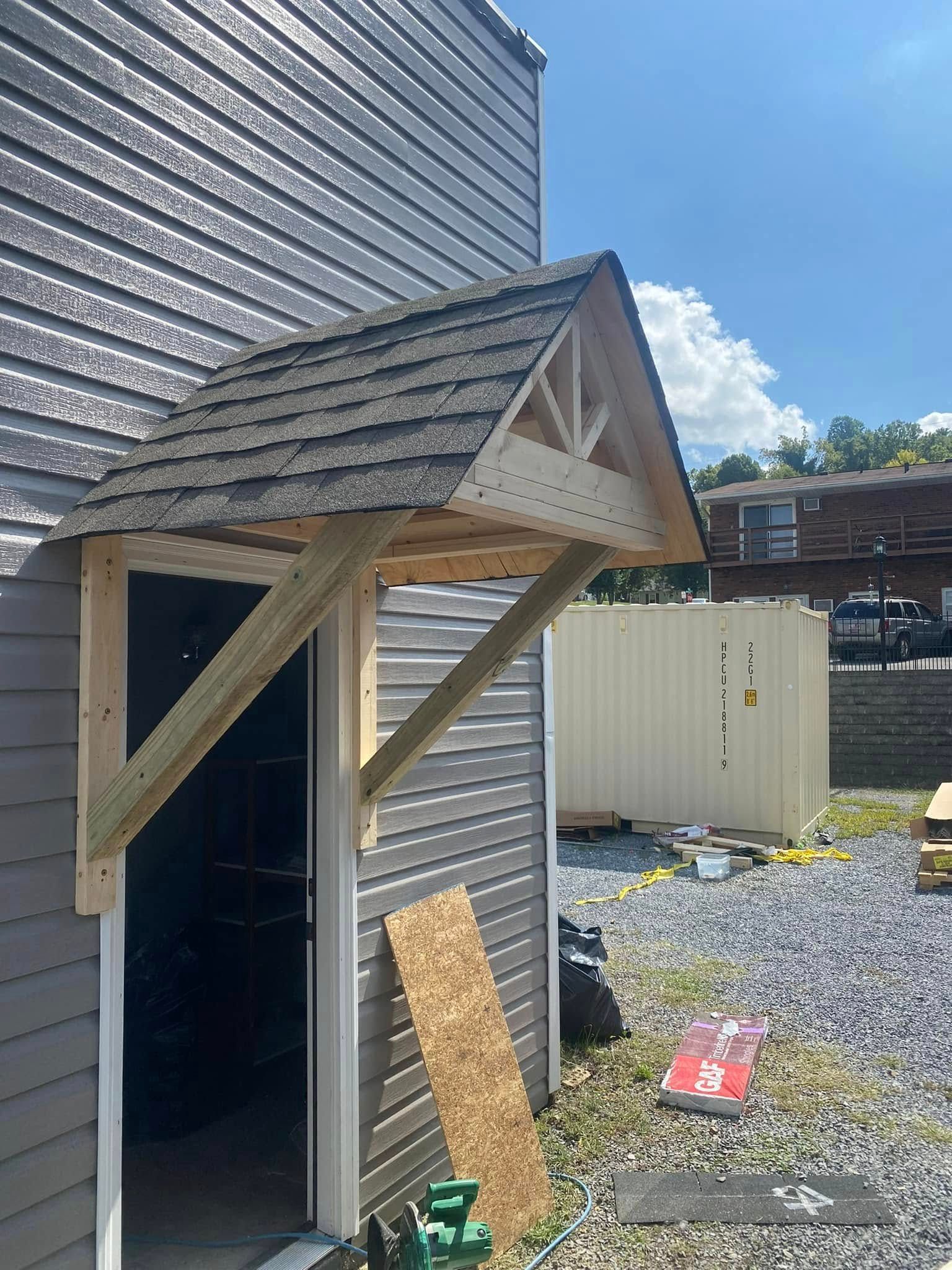 A newly constructed wooden door overhang with asphalt shingles attached to the gray siding of a building exterior.