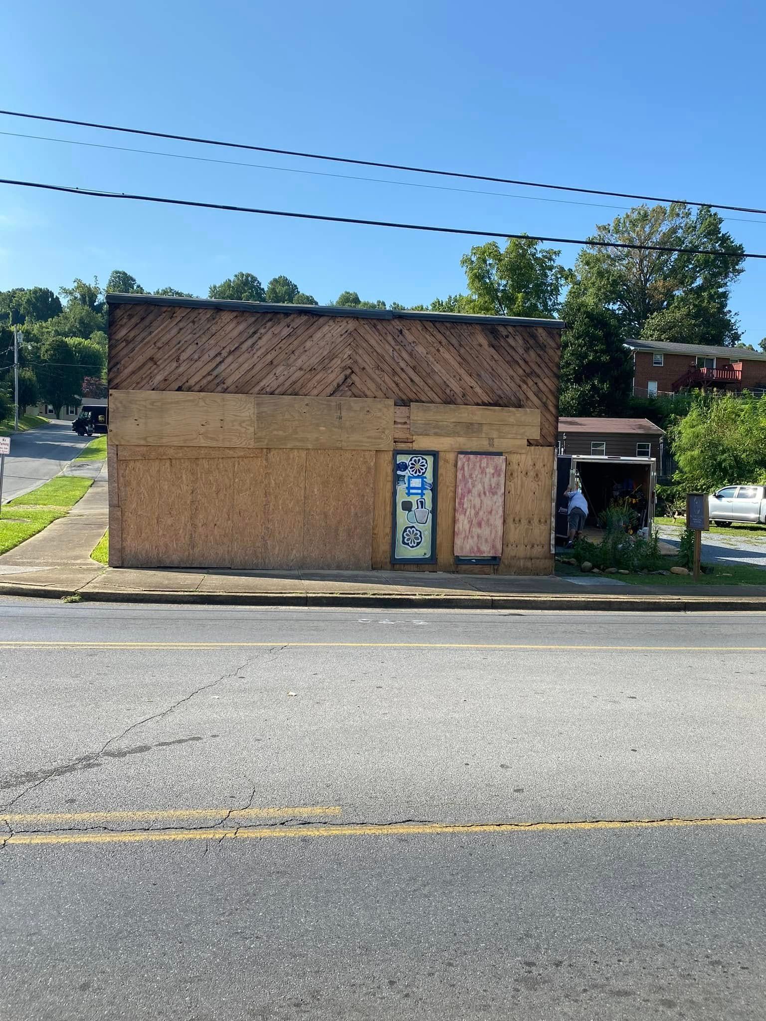 A small, boarded-up commercial building with graffiti on the door, standing along a sunny, paved street.
