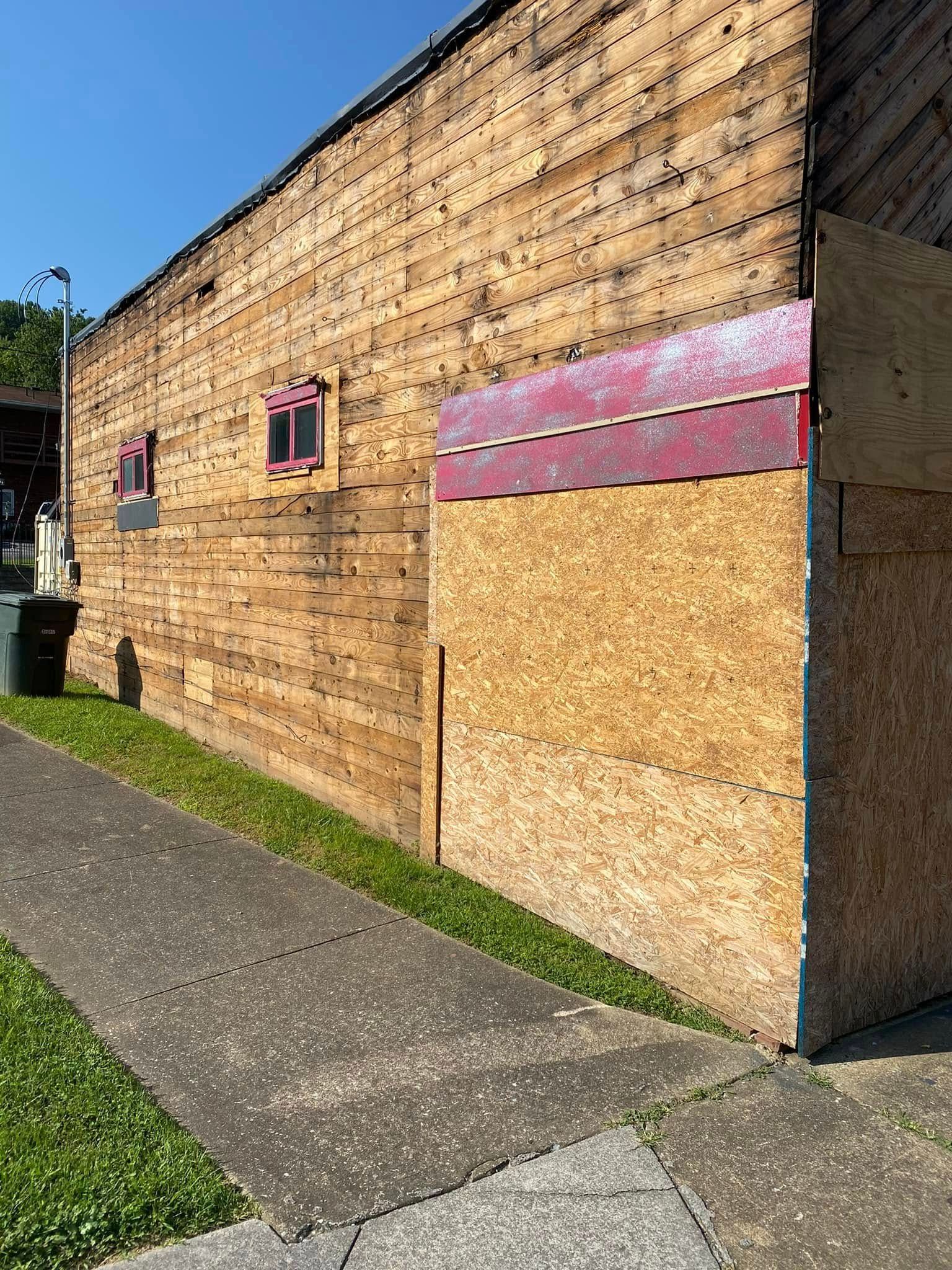 A weathered wooden building exterior featuring a section covered in plywood, with small windows and a sidewalk.