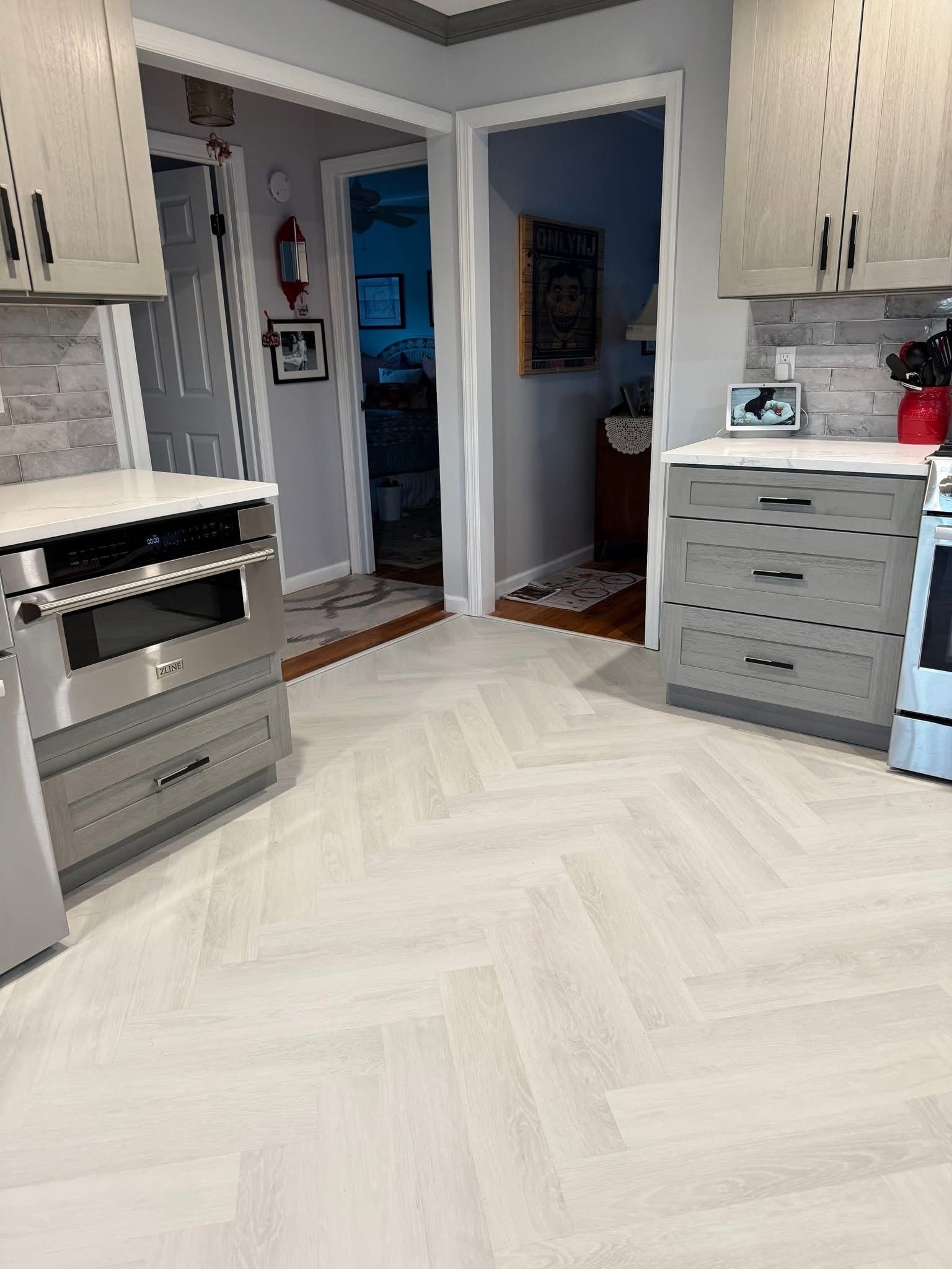Modern kitchen with white cabinets, herringbone floor, and a doorway to a dark room