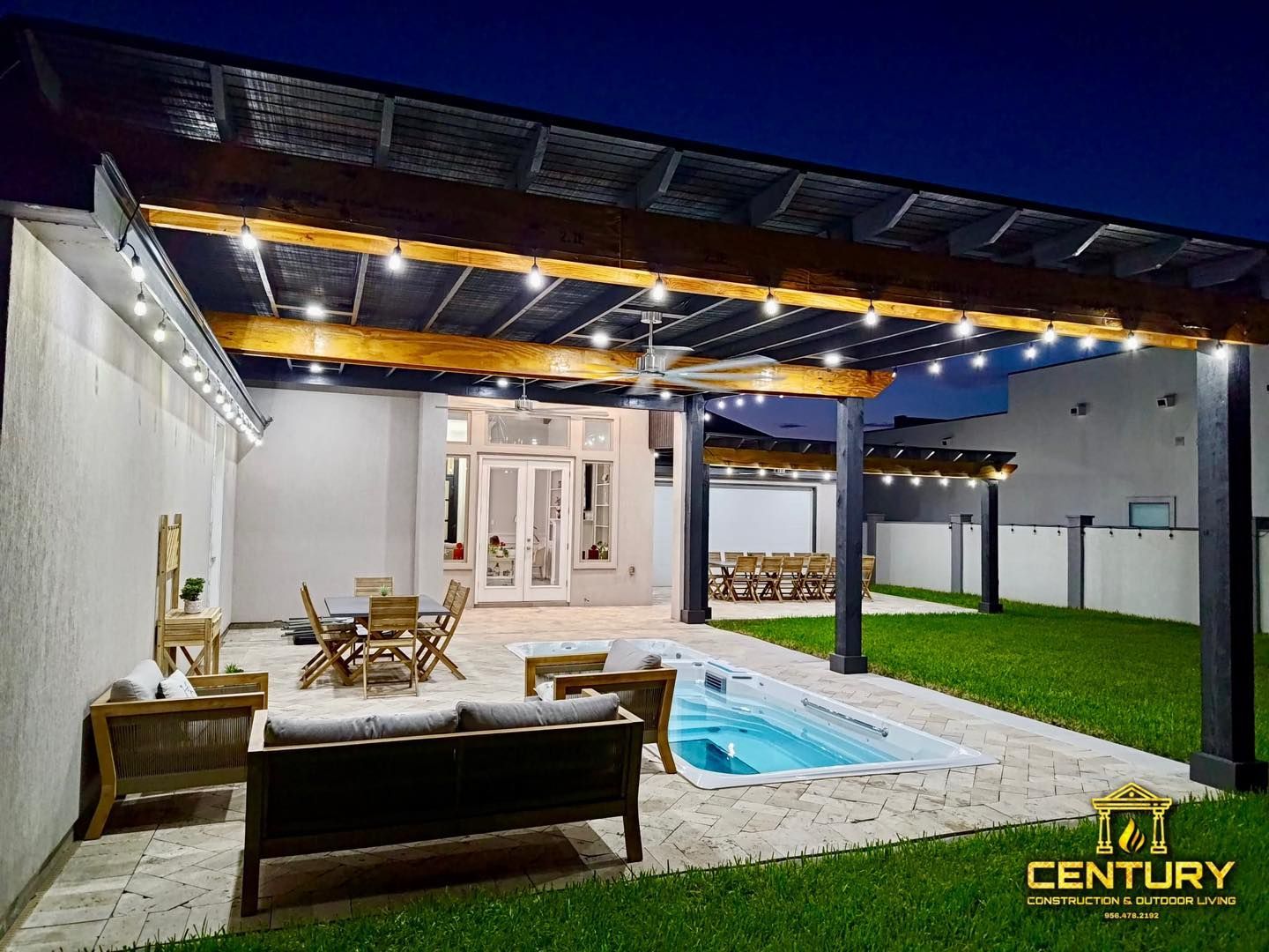 A patio with a swimming pool and a pergola at night.